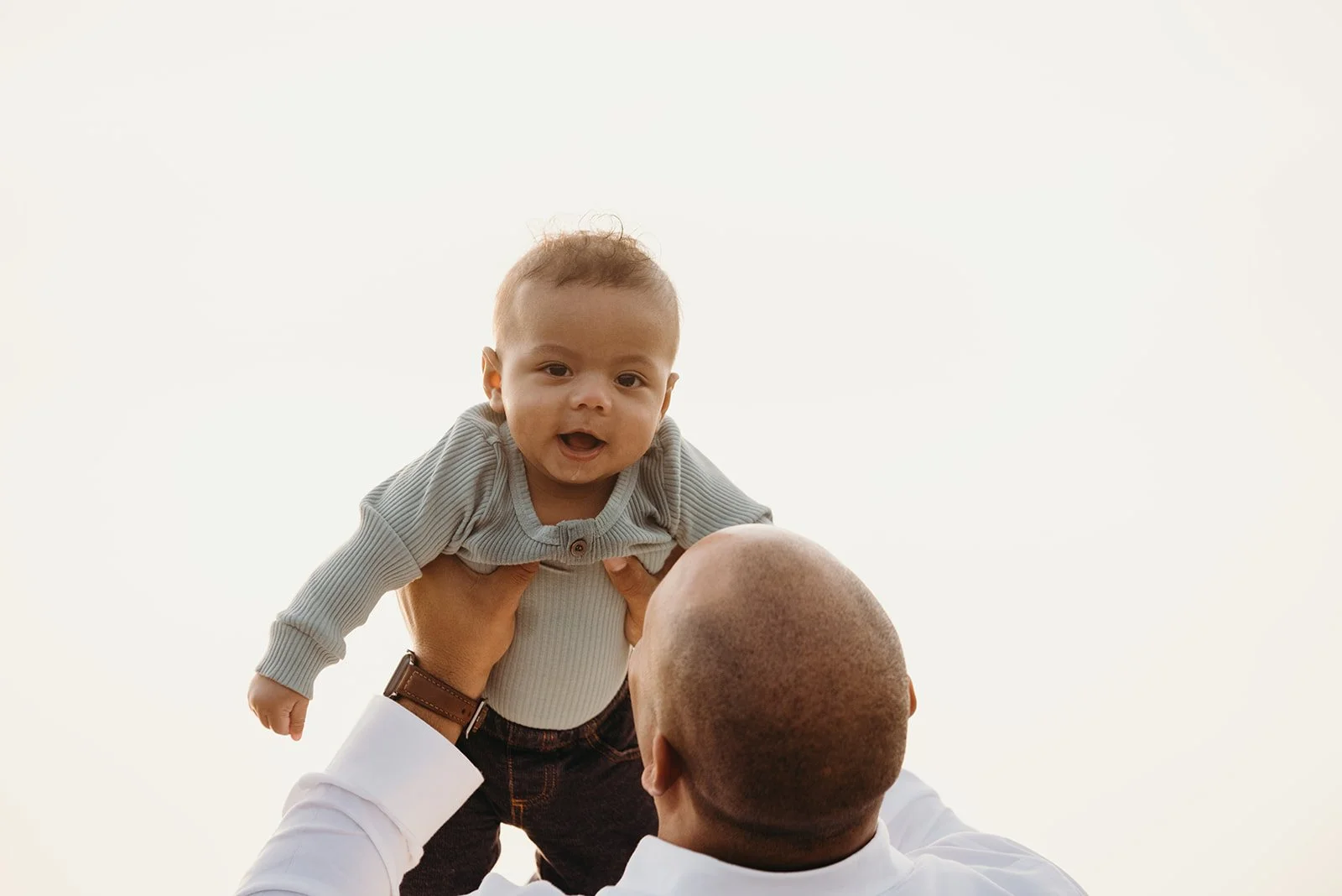 A man holding up a smiling baby in the air against a bright background.