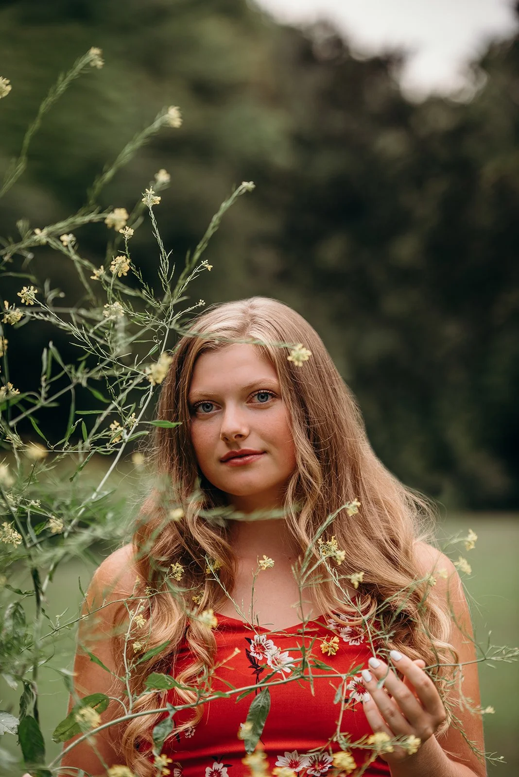 A young woman with long, wavy hair, wearing a red floral dress, standing outdoors surrounded by green foliage and plants.