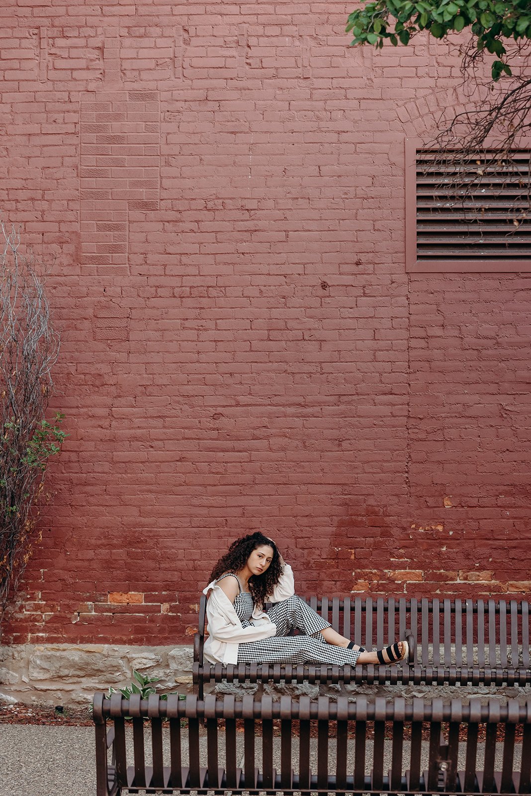A woman with curly hair sitting on a bench against a red brick wall. She is wearing a black and white checkered jumpsuit, a white jacket, and sandals, posing with one leg extended and one arm resting on her head.