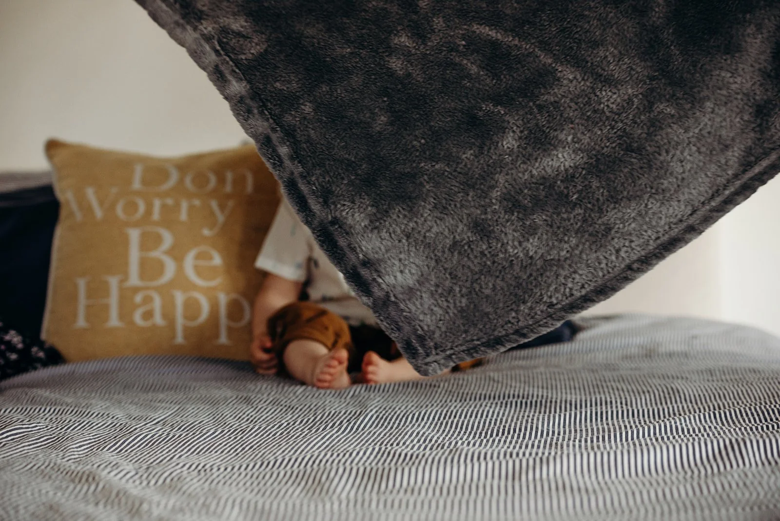 A bed with striped bedsheets, a large gray pillow hanging over the bed, a brown pillow with white text "Don’t Worry Be Happy," and a person sitting on the bed with only their legs and part of their arm visible, holding a small brown stuffed animal.
