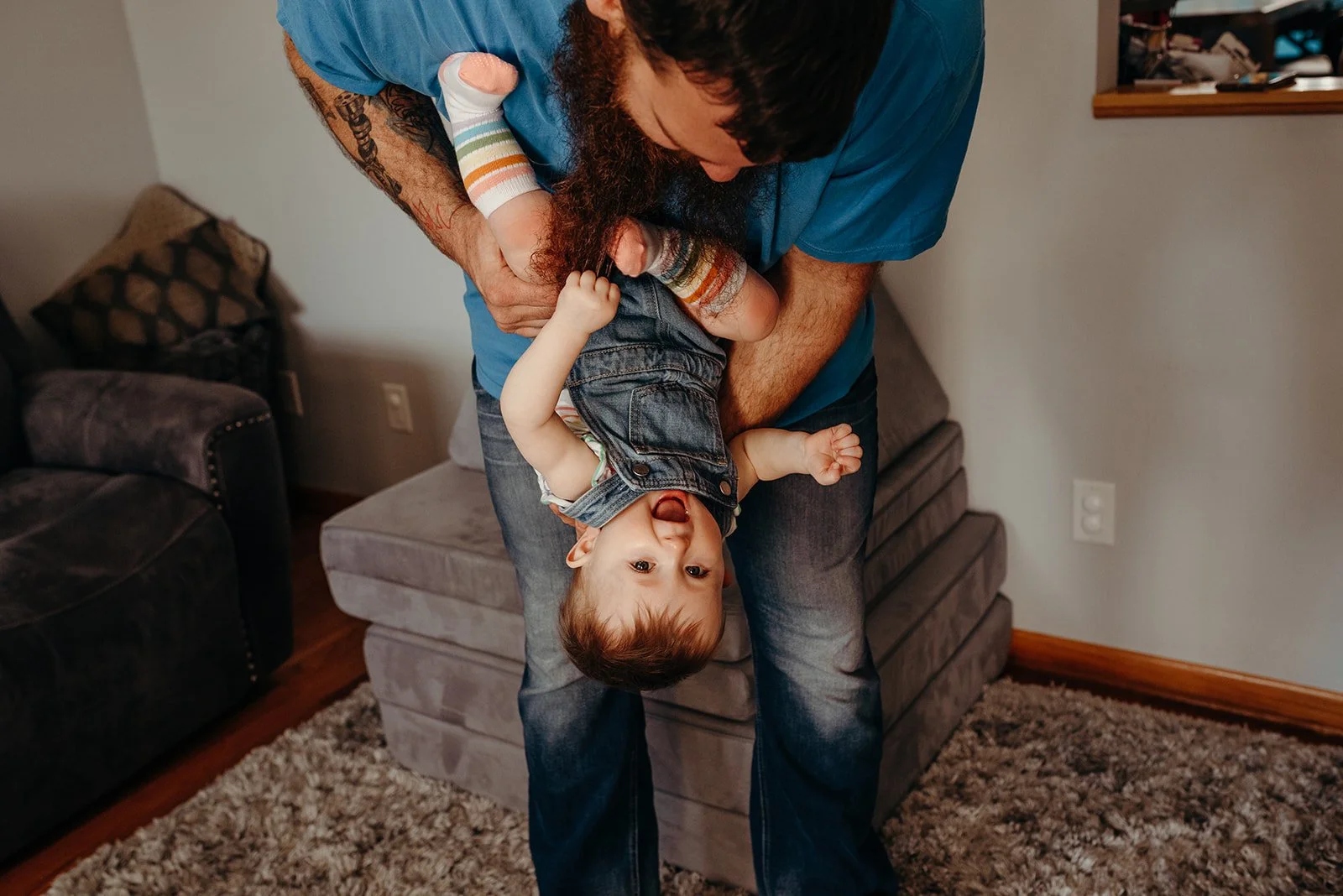 A man is holding a laughing toddler upside down, lifting him by his waist, inside a living room with a rug, couch, and a small table.