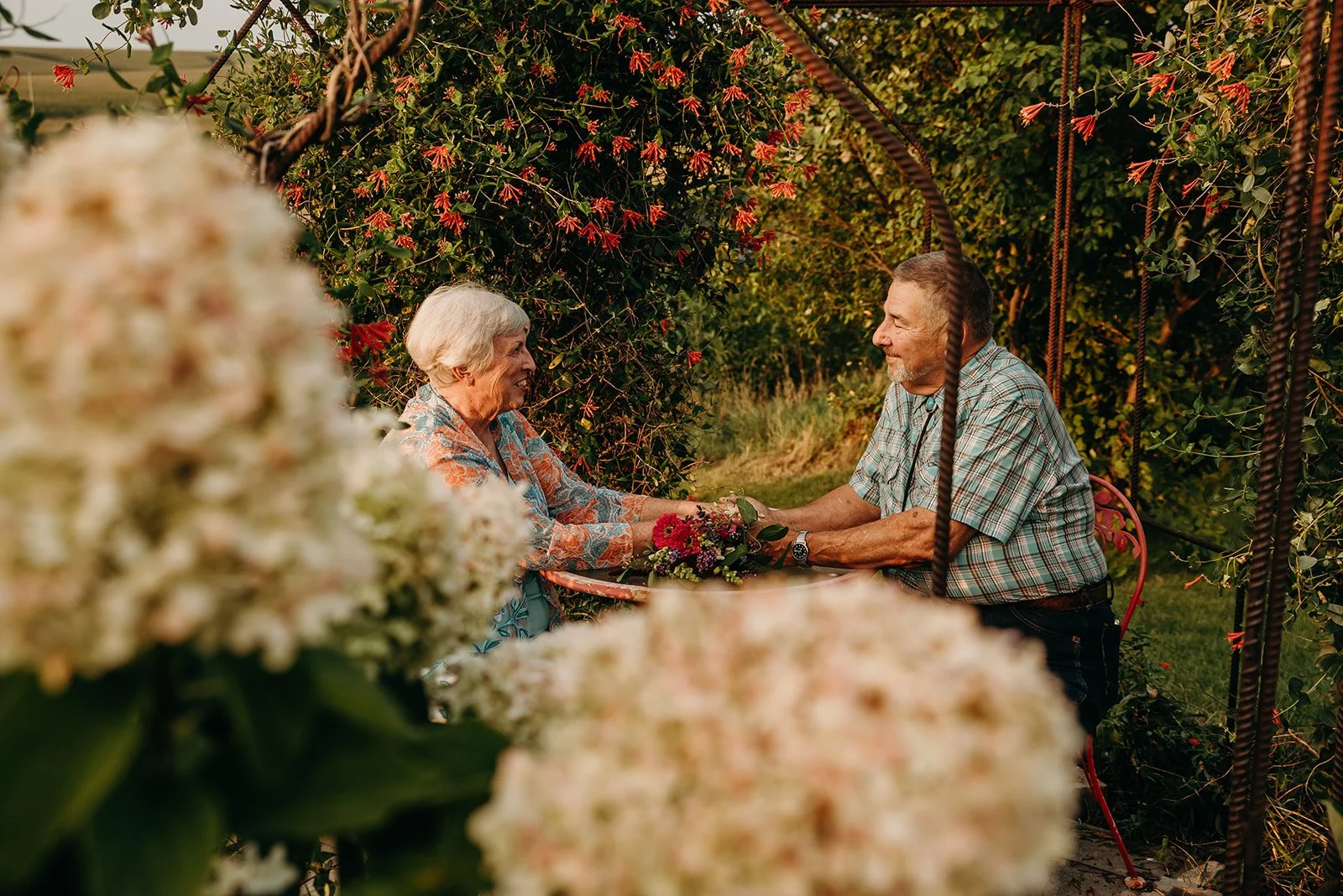 An elderly couple sitting on a swing in a lush garden, holding hands and smiling at each other with a bouquet of flowers on the table.