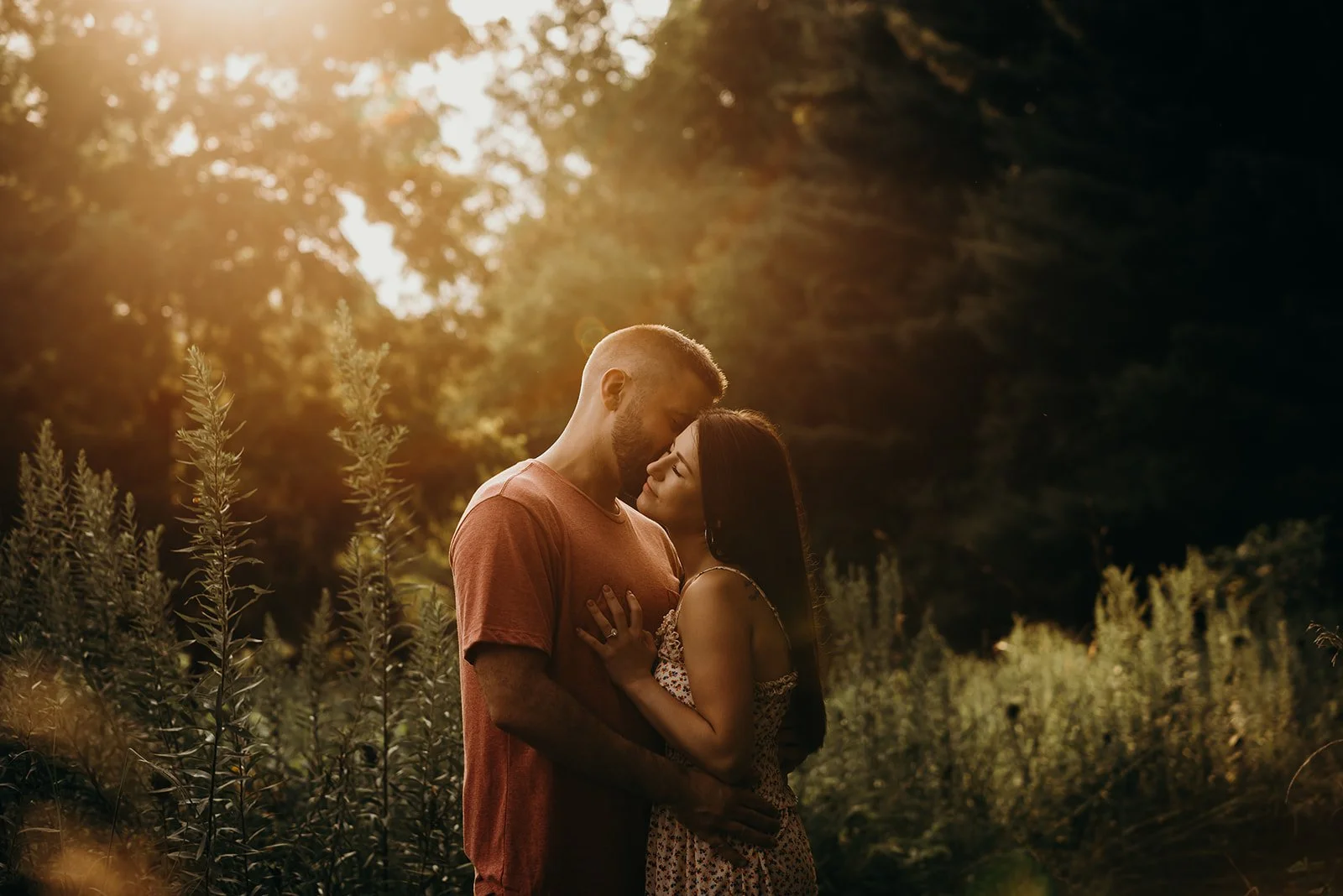 A couple embracing outdoors during sunset in a natural setting with trees and tall grasses.