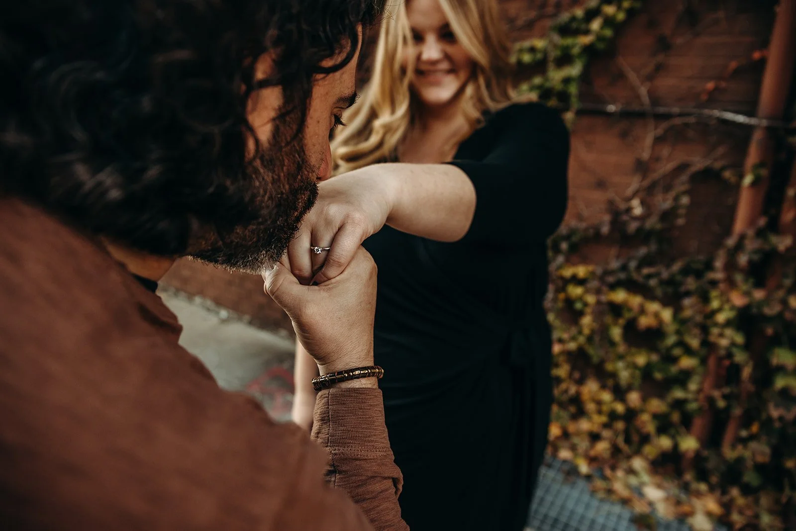 A man is kissing a woman's hand outdoors. The woman is smiling and has blonde hair. They are near a wooden fence with plants and foliage.