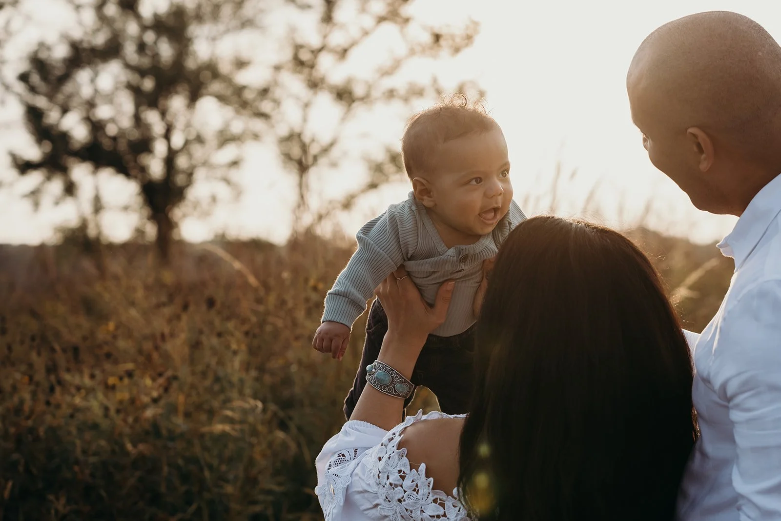 A happy family outdoors at sunset, with a woman holding a smiling baby in the air, while a man looks on.