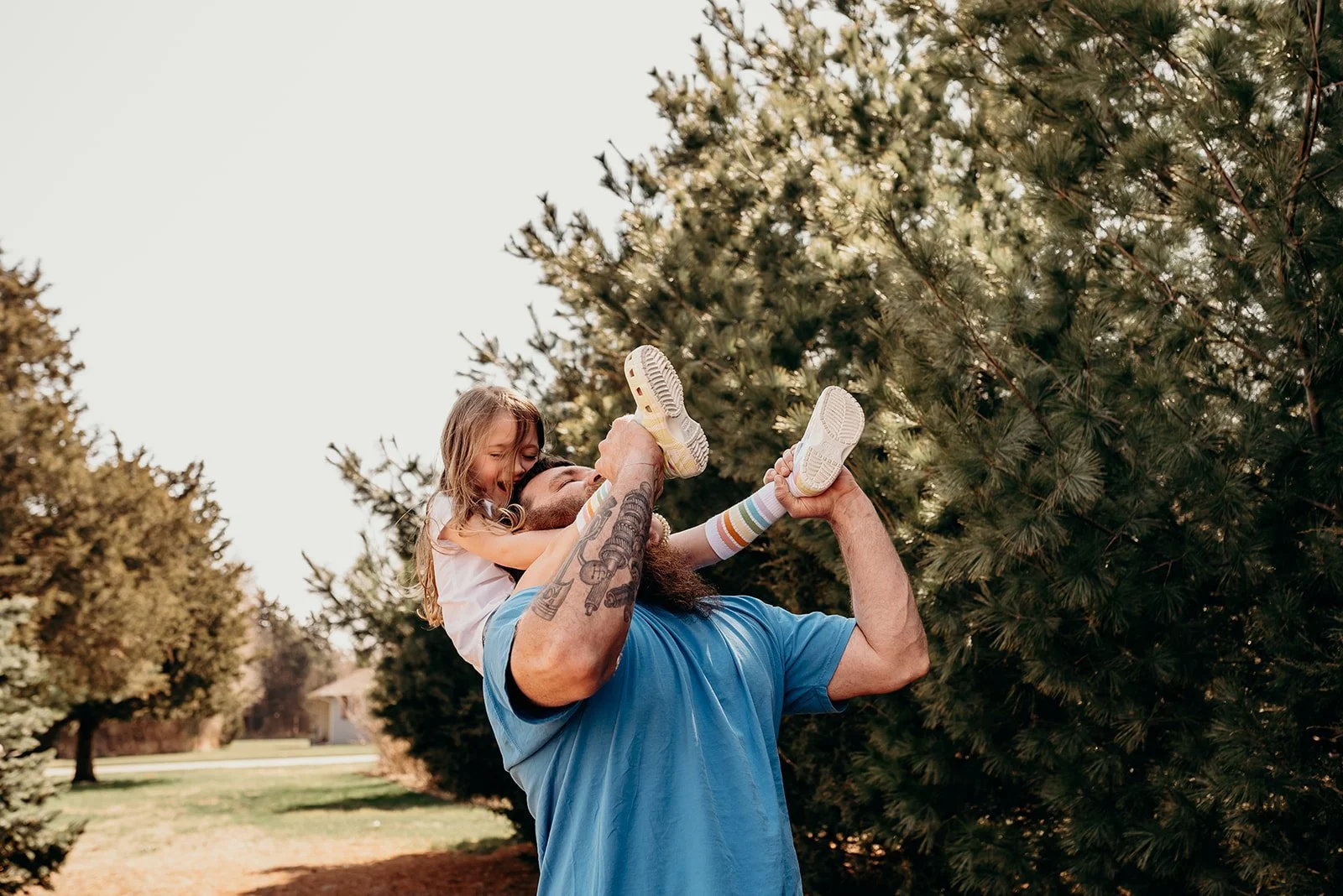 A man with tattoos lifting a young girl with long hair into the air outdoors with trees in the background.