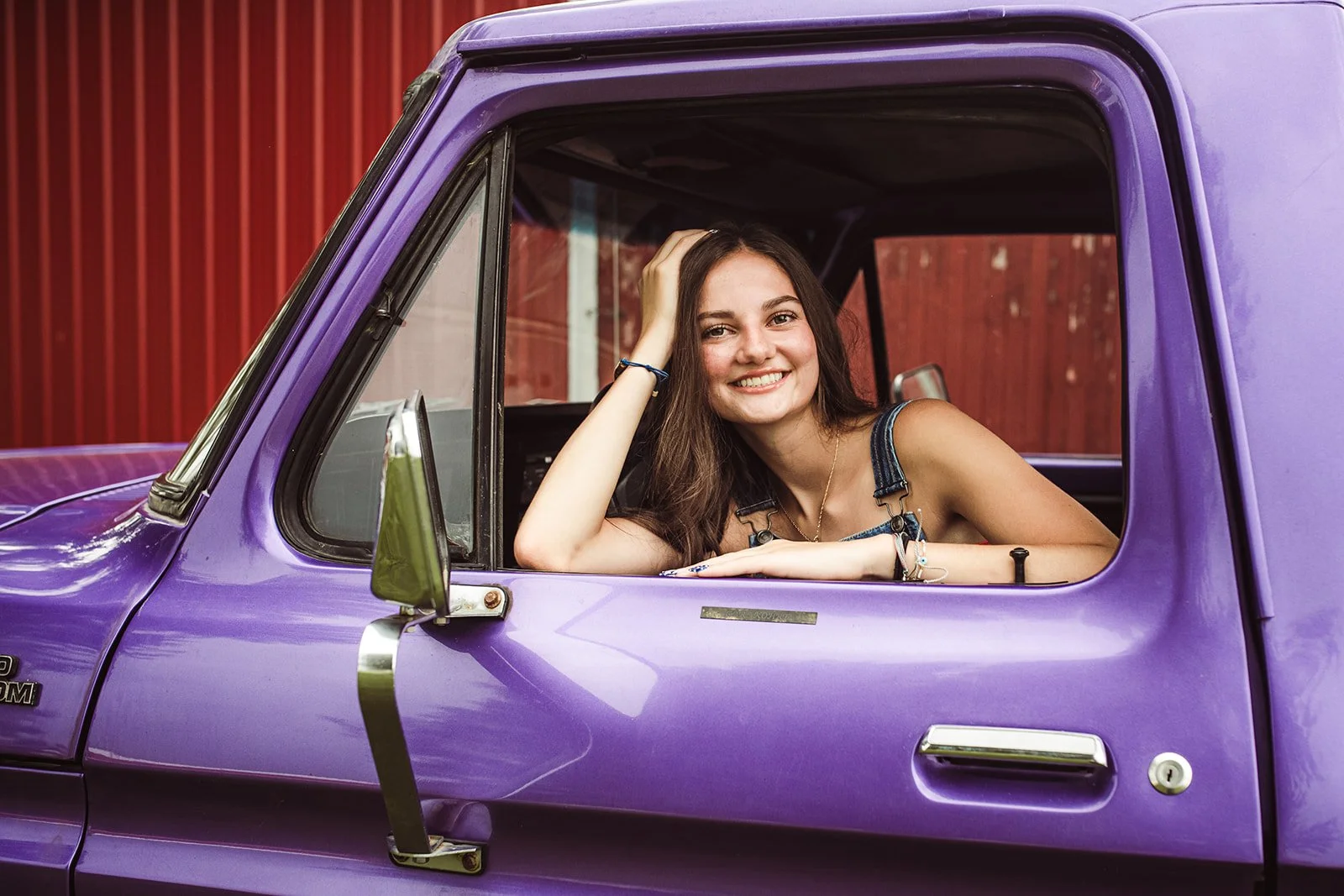 A smiling young woman with long brown hair sitting in the driver's seat of a purple pickup truck, resting her arm on the window frame with her hand on her head.