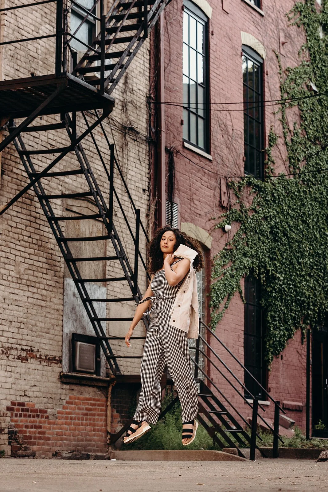 A woman with curly hair wearing checkered jumpsuit, sneakers, and a beige jacket draped over her shoulder, standing in an urban alleyway with brick buildings and fire escape stairs.