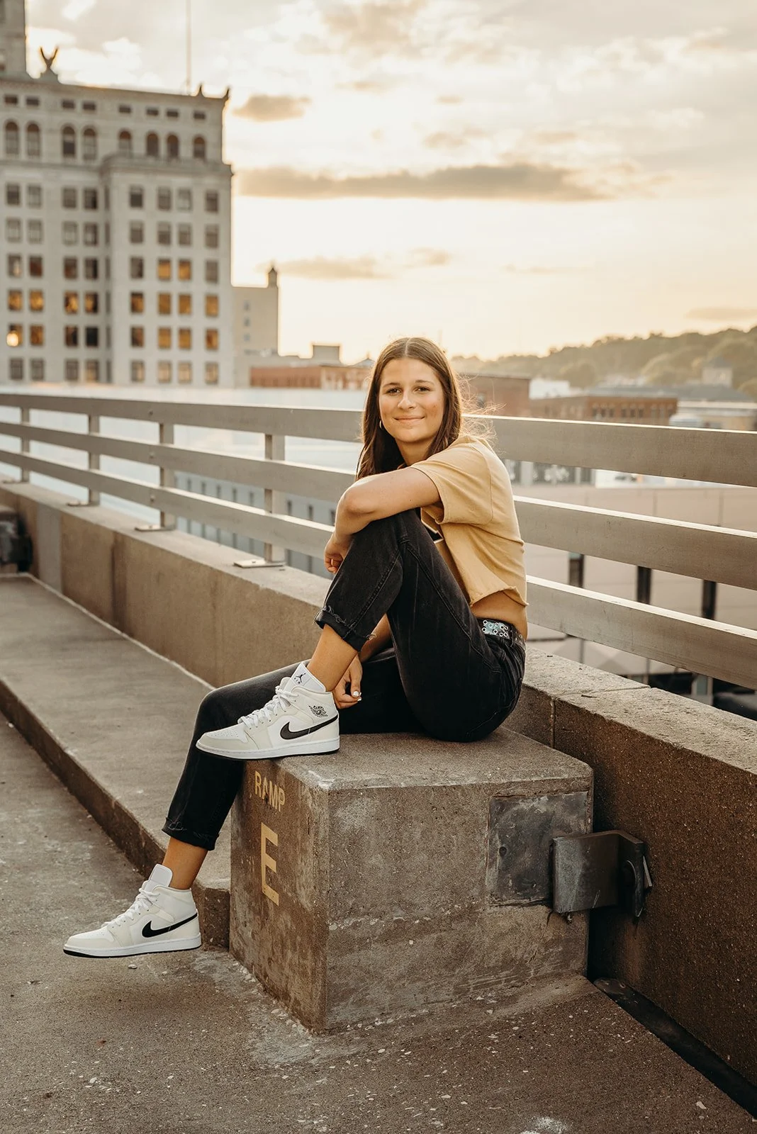 A young woman sitting on a concrete ledge on a rooftop parking structure during sunset, smiling at the camera, wearing a beige t-shirt, black jeans, and white Nike sneakers.