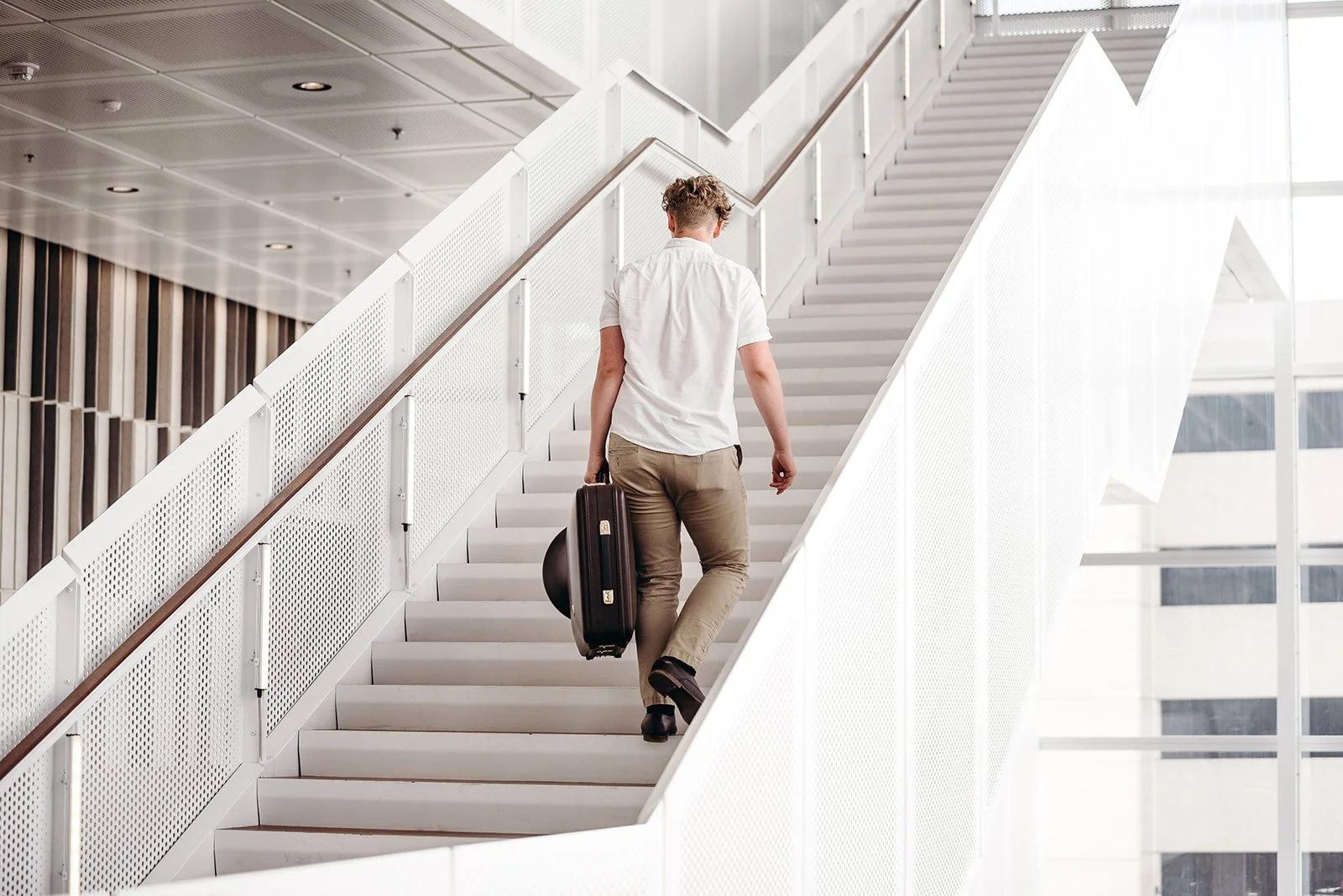 A young man with sunglasses, a white shirt, beige pants, and black shoes walking up a modern white staircase with a rolling suitcase.