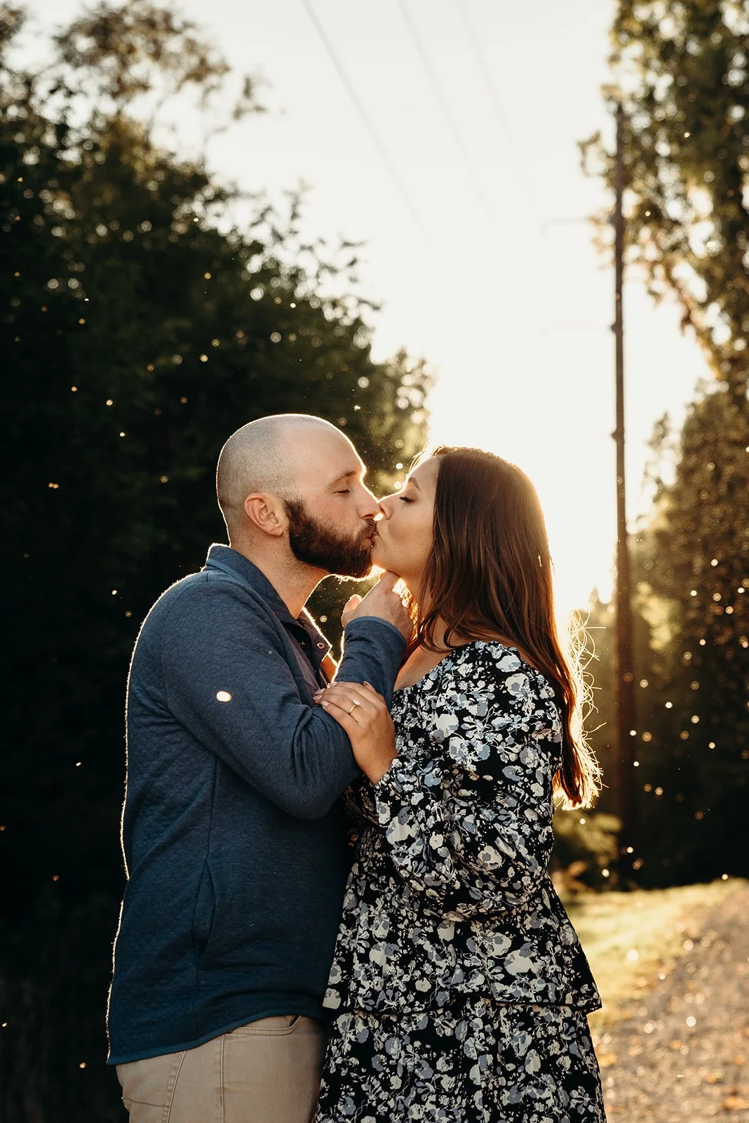 A couple sharing a kiss outdoors in the sunset, with trees and power lines in the background.