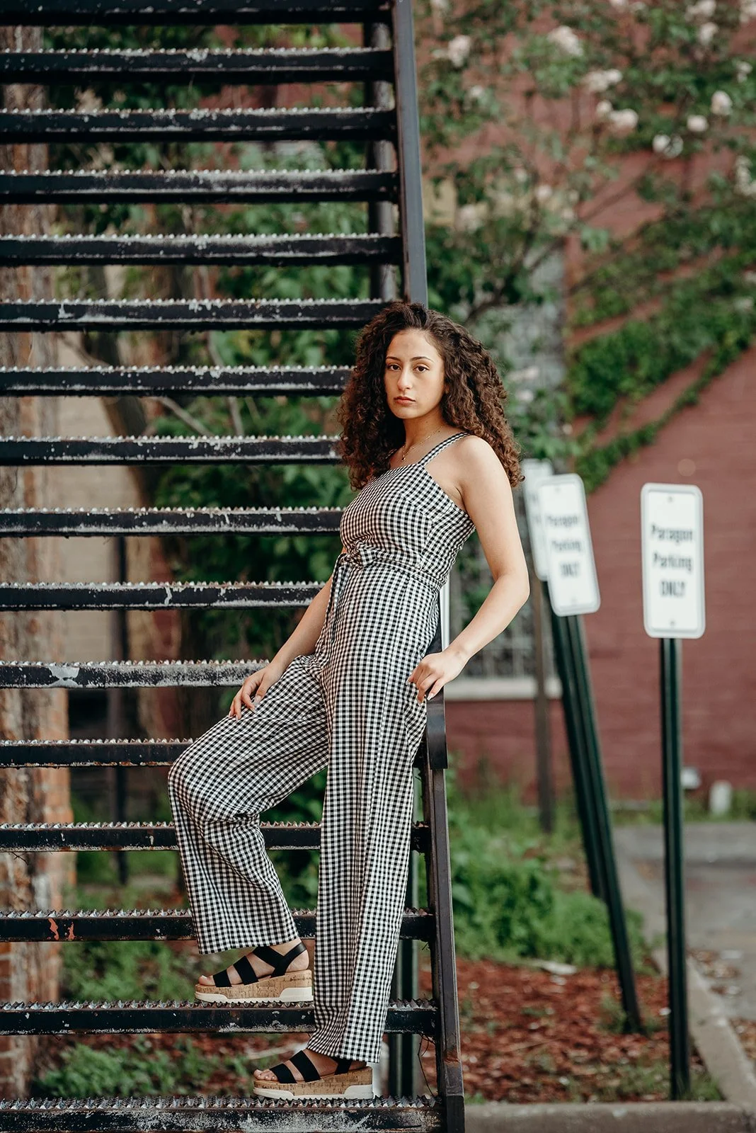 A young woman with curly hair wearing a black and white checkered jumpsuit standing next to an outdoor staircase with snow or ice on the steps, in an urban setting with greenery and parking signs in the background.