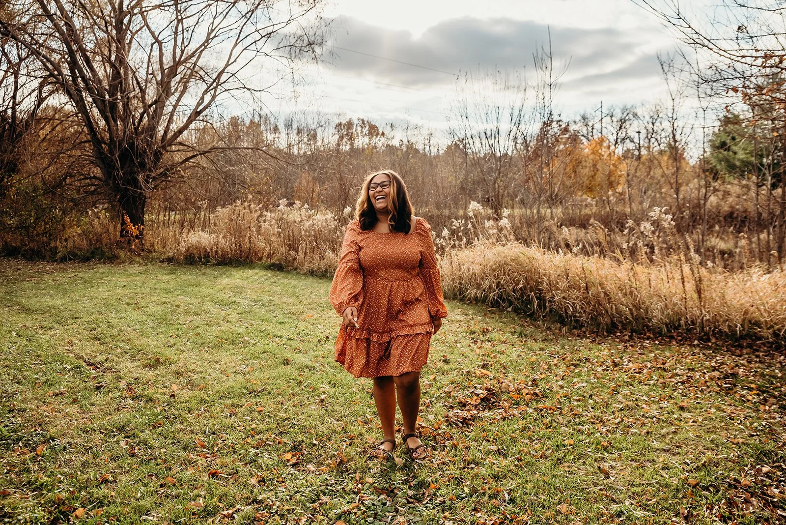 A woman in a rust-colored dress with puffed sleeves, walking on a grassy field with autumn leaves, laughing, and holding a phone, with trees and cloudy sky in the background.