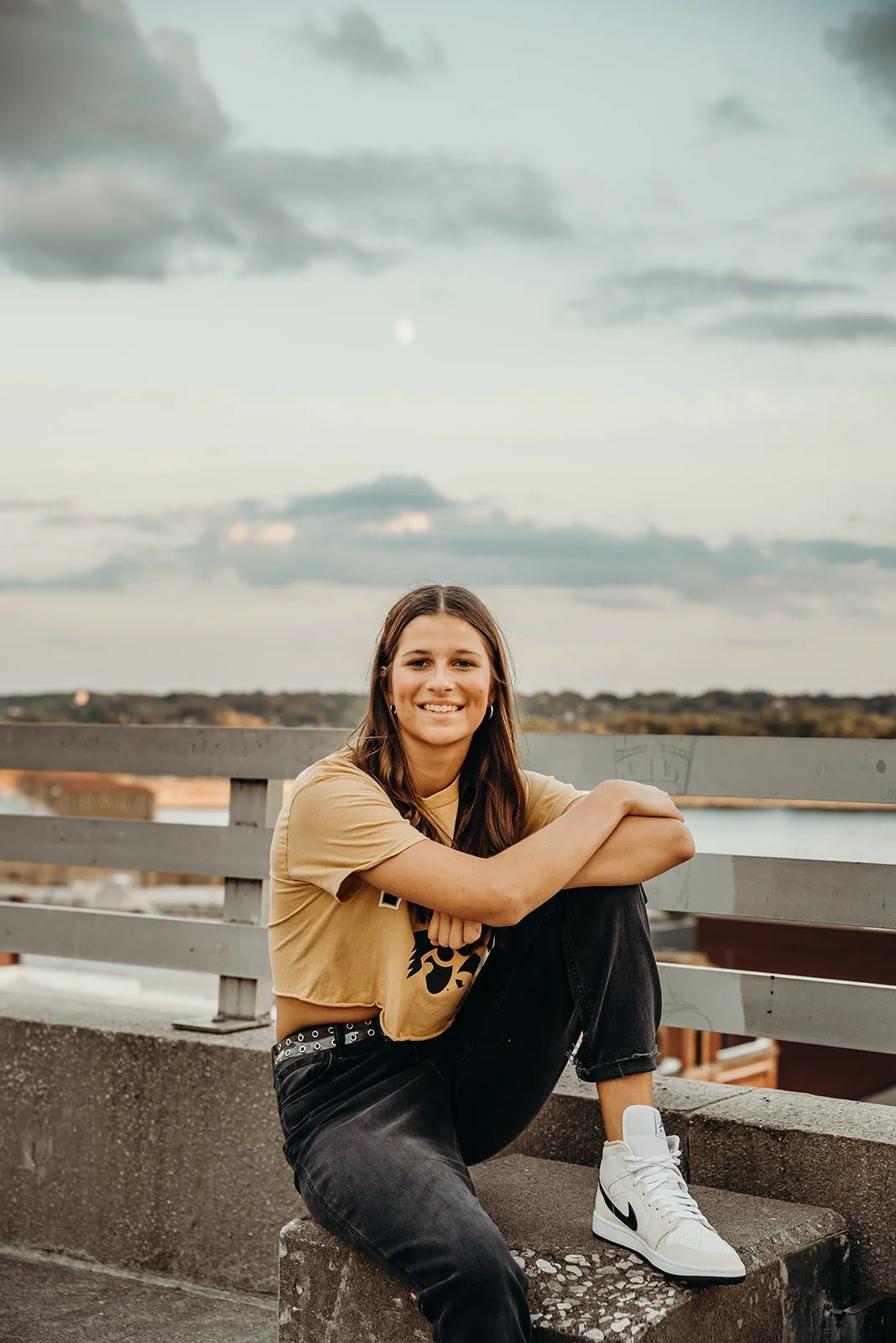 A woman sitting on a concrete ledge outdoors during the evening, smiling at the camera, with a body of water and cloudy sky in the background.