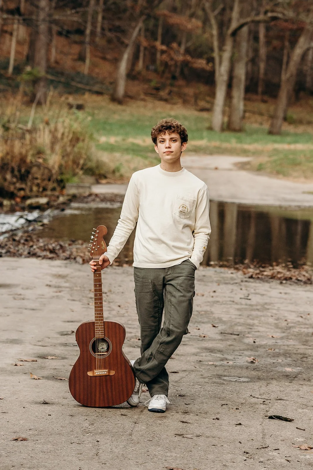 Young man standing outdoors with a guitar, near a creek, during fall.