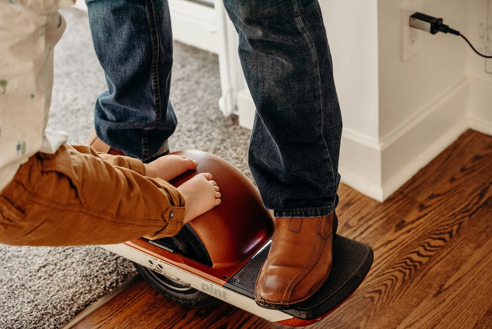 Person standing on a hoverboard while holding a round object, possibly a helmet, near another person in a room with hardwood flooring and a carpet.