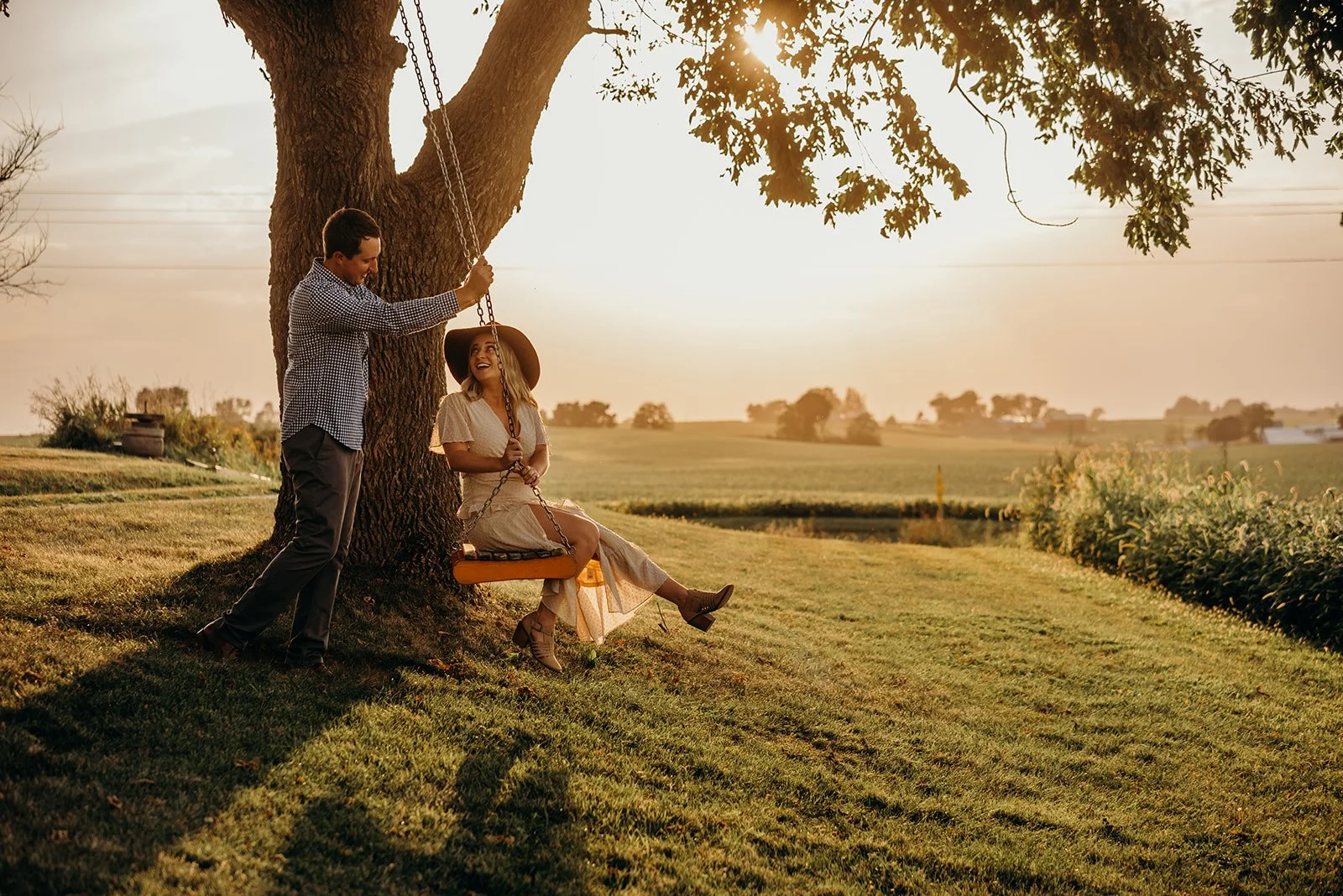 A man and woman enjoying a sunset under a large tree in a rural setting; the woman sits on a swing, wearing a dress and sunhat, while the man stands next to her holding the swing's ropes, smiling at her.