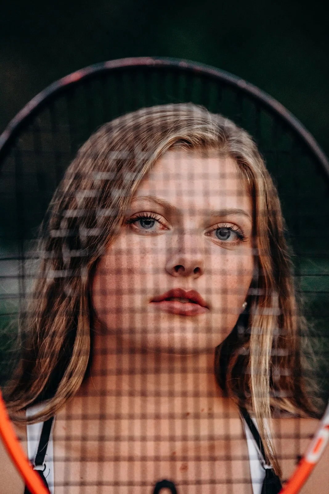 Close-up of a young woman with blonde hair and blue eyes, looking at the camera through a tennis racket with smudged lines on the glass.