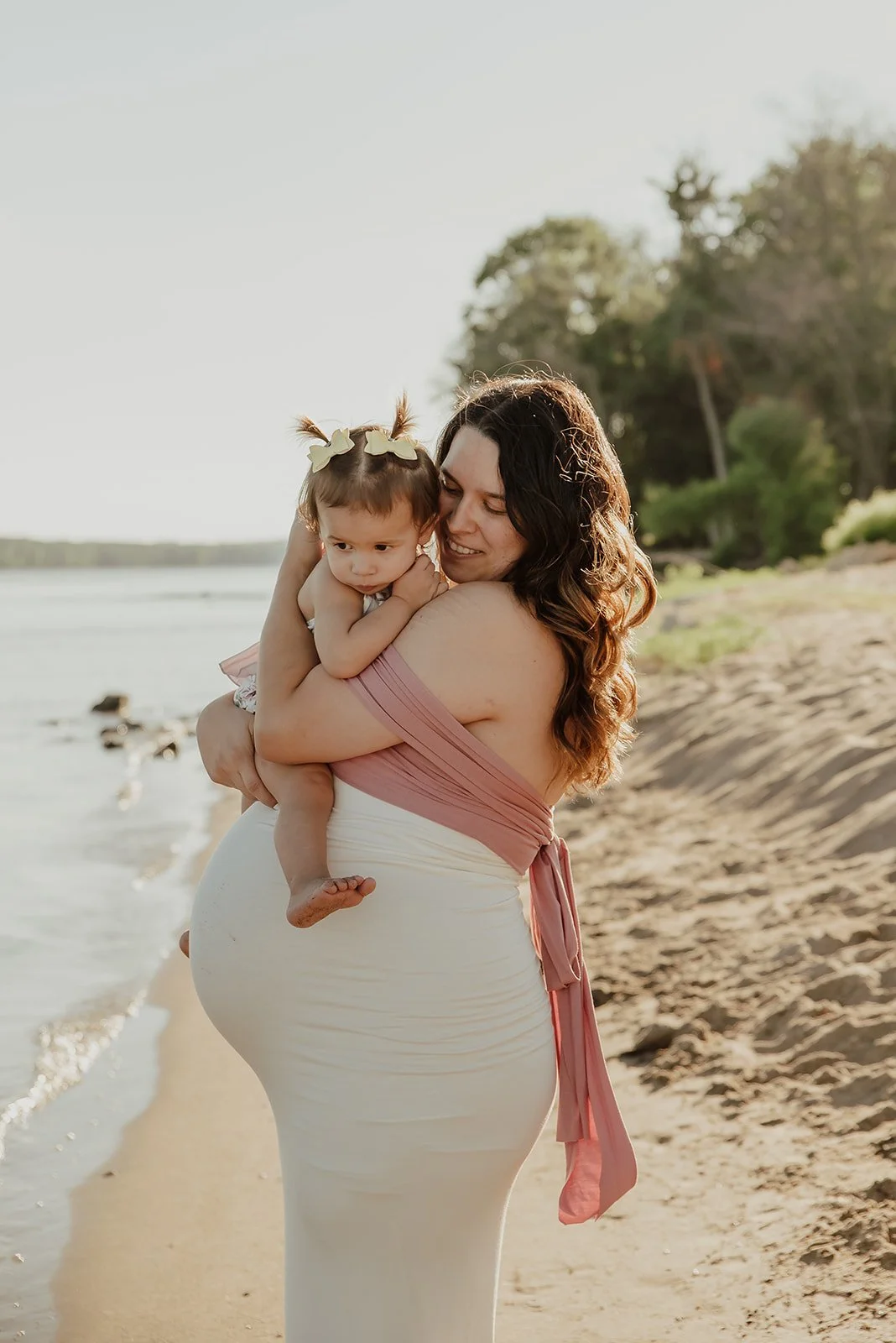 A woman holding a young girl on a beach during sunset, with trees in the background.