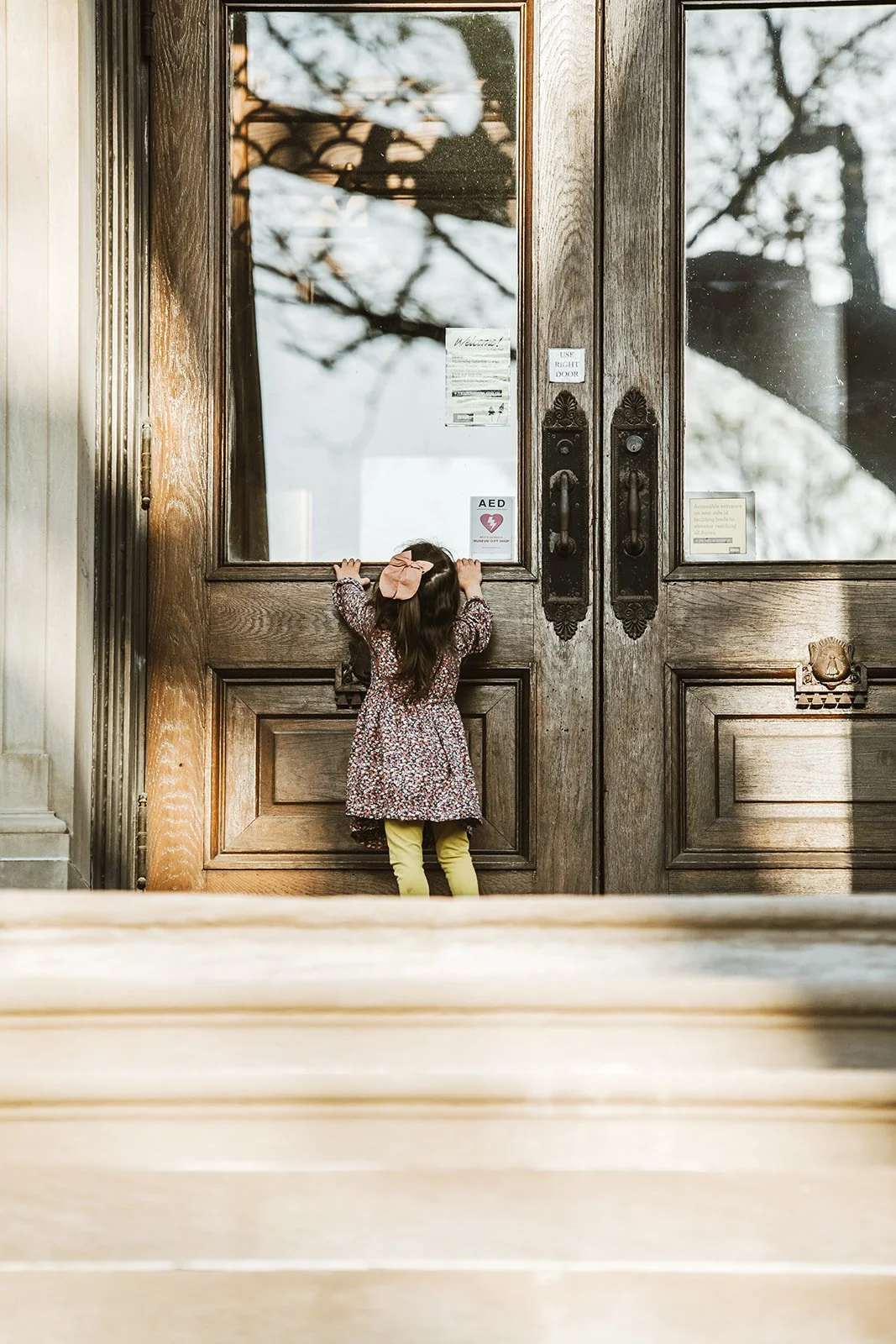 A young girl standing outside a large wooden door holding onto the door with her hands, looking inside through the glass panel.