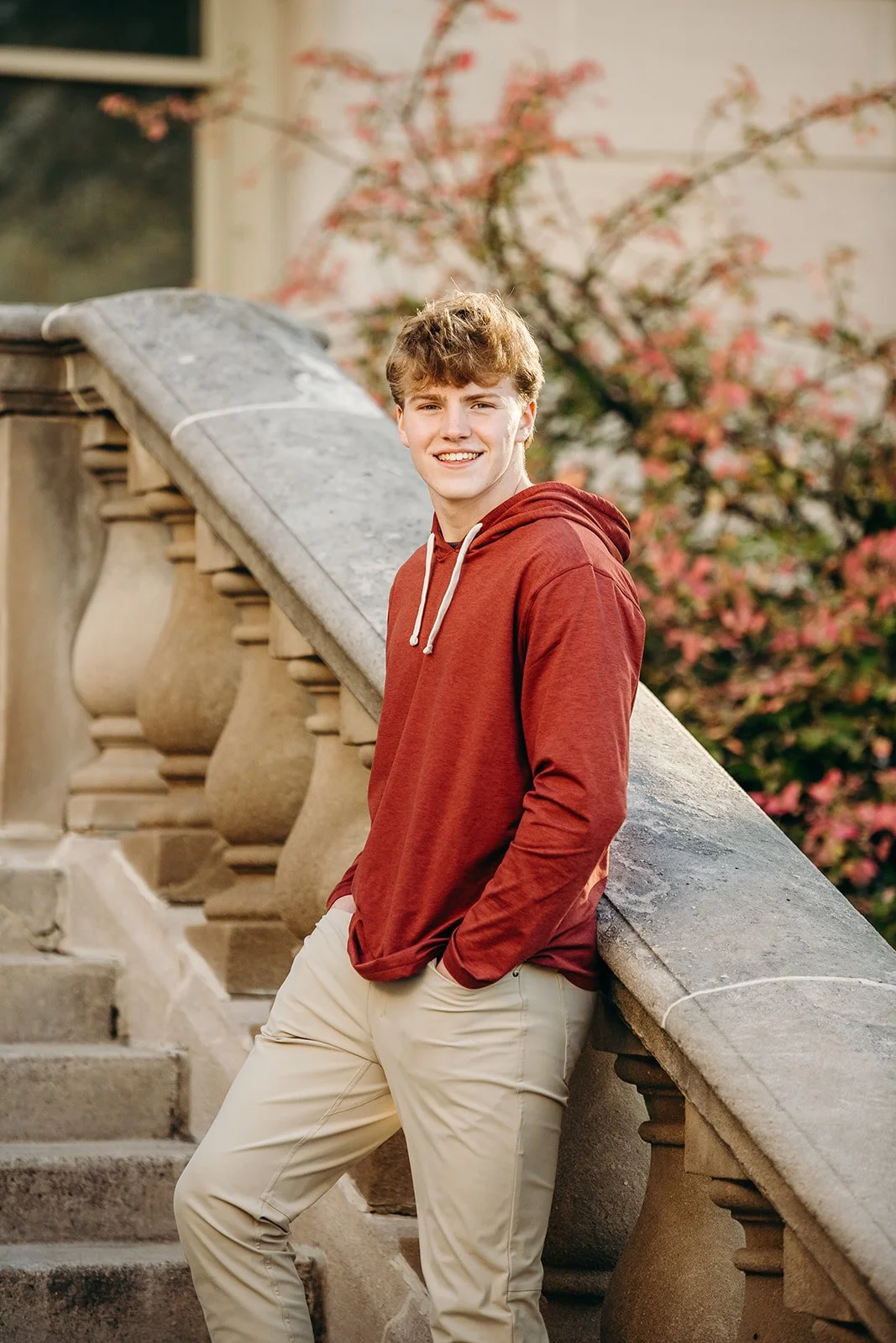 A young man with curly blond hair smiling, wearing a red hoodie and beige pants, leaning against a stone railing on outdoor stairs with a blooming pink tree in the background.