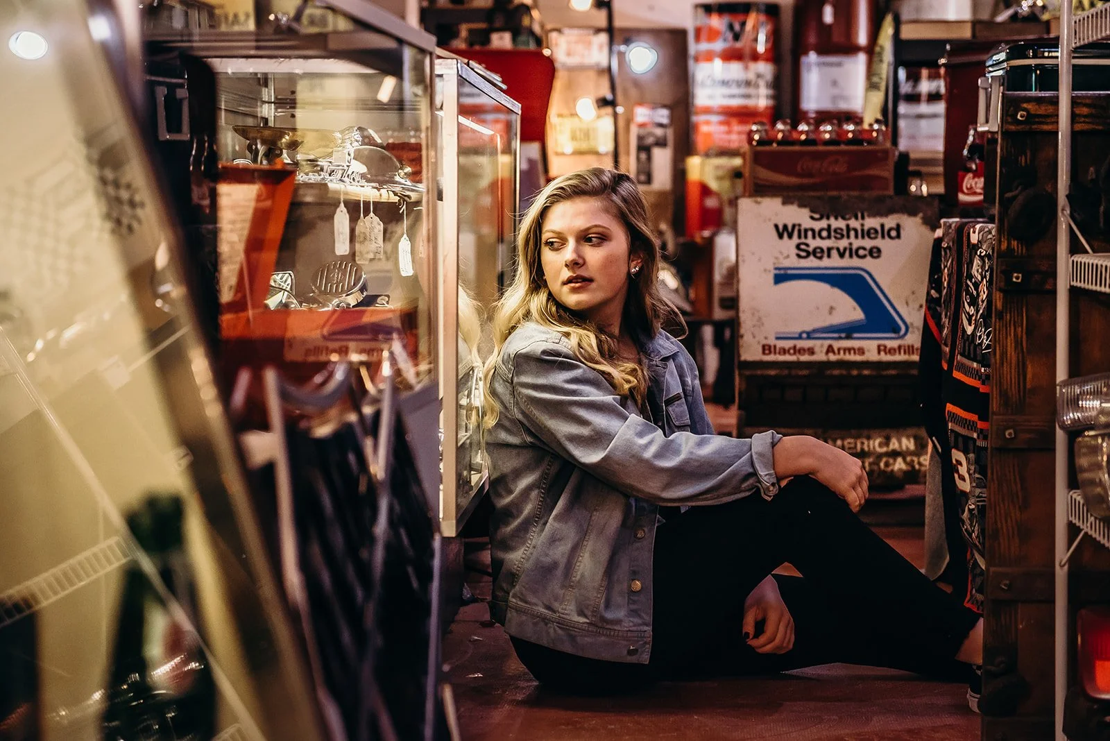 A young woman with blonde wavy hair wearing a gray jacket sitting on the floor of a store aisle, surrounded by shelves of kitchen utensils and other items, looking to her side with a serious expression.
