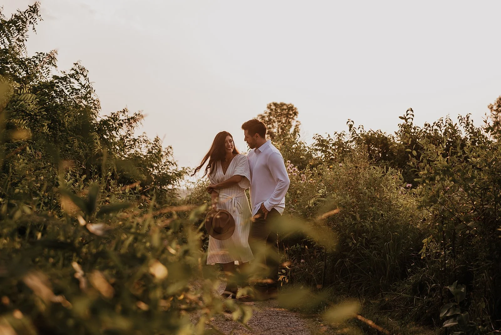 A young couple walking and smiling in a lush garden during sunset, with greenery and blooming plants around them.