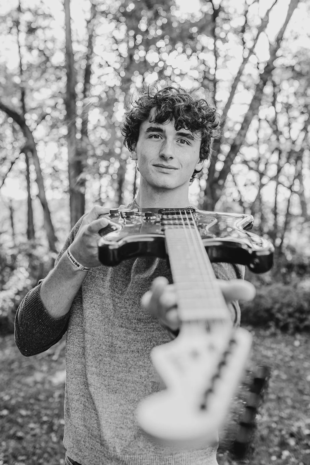 A young man with curly hair holding a guitar and looking at the camera, outdoors among trees, in black and white.