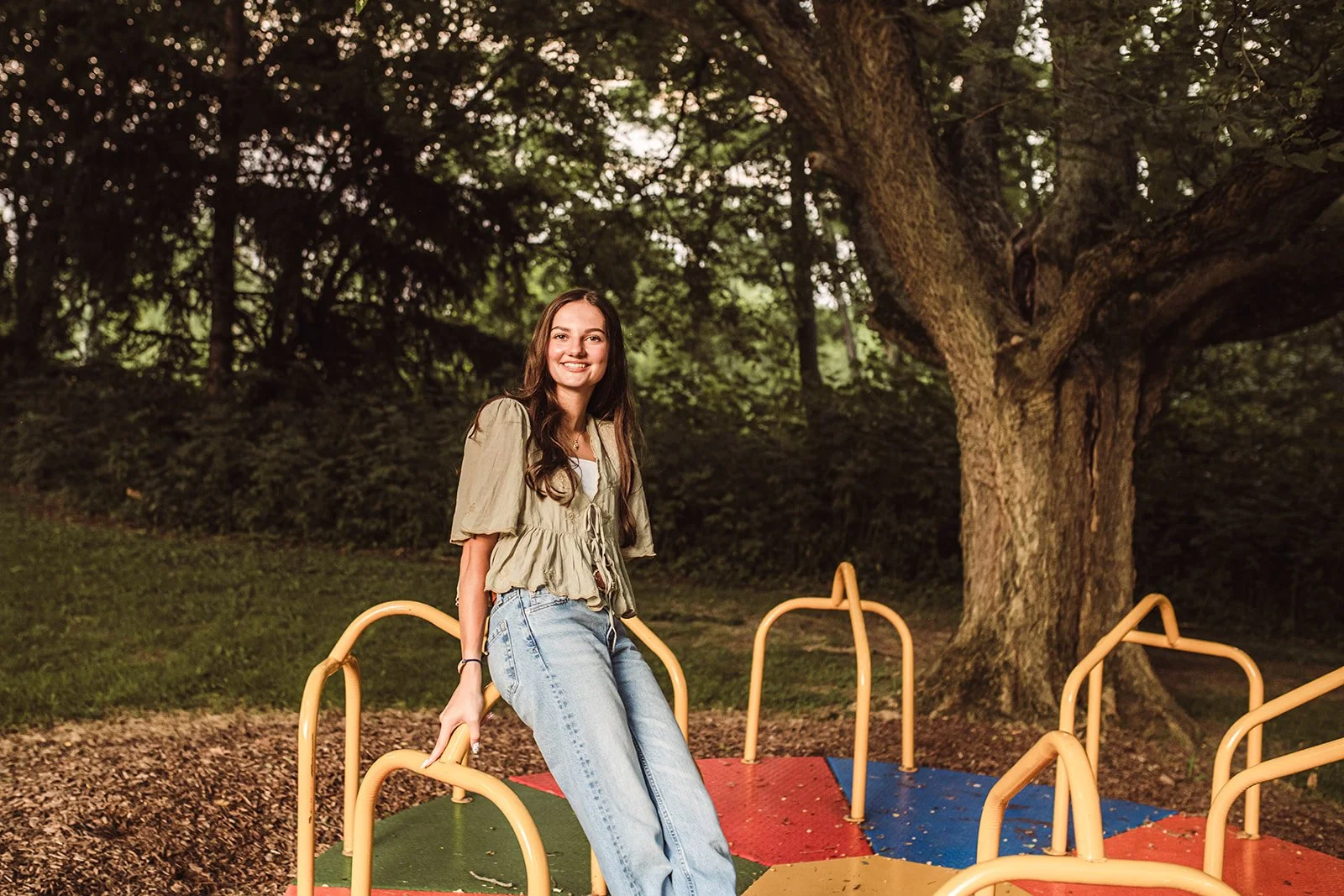 A young woman with long brown hair sitting on a colorful merry-go-round in a park with trees and green foliage in the background, smiling at the camera.