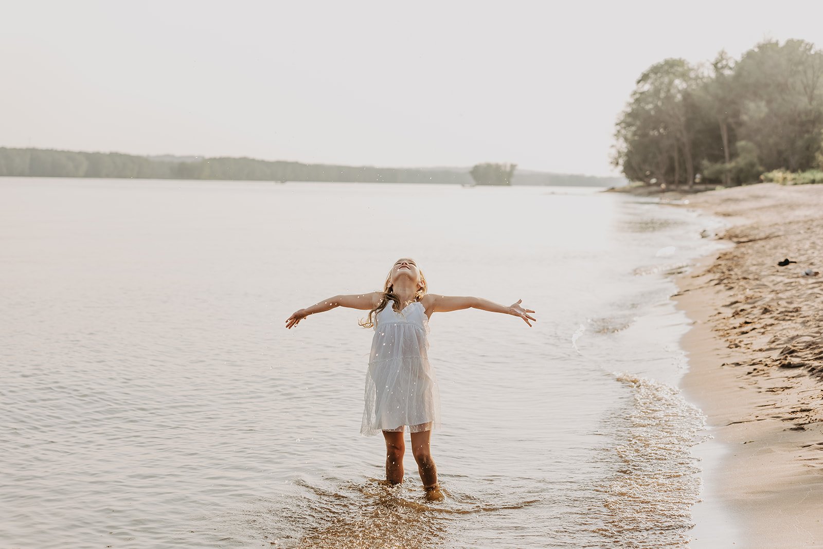 A young girl in a white dress stands in shallow water with arms outstretched, enjoying a beach scene with a sandy shoreline and trees in the background.