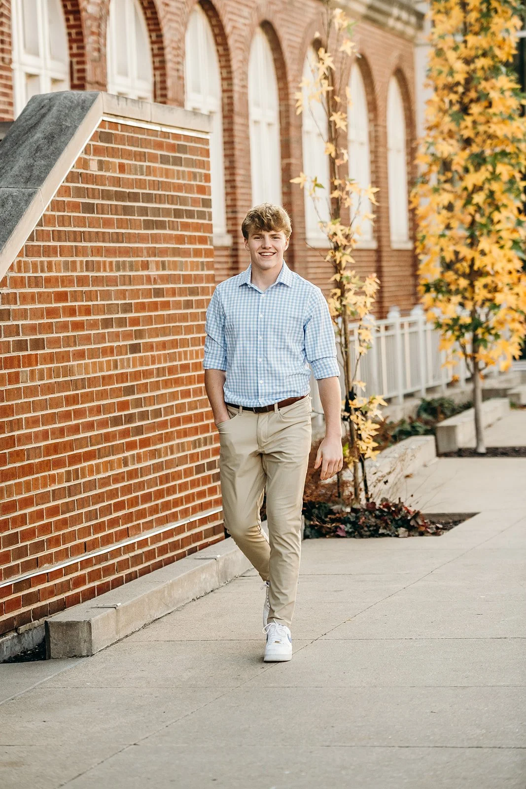 A young man with light brown hair in a checkered shirt and khaki pants walking on a sidewalk lined with trees that have yellow and orange leaves, in front of a historic brick building.