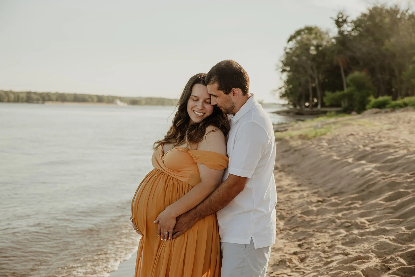 A pregnant woman in a flowing orange dress and a man in a white shirt sharing a tender moment on a sandy beach by the water, surrounded by trees.