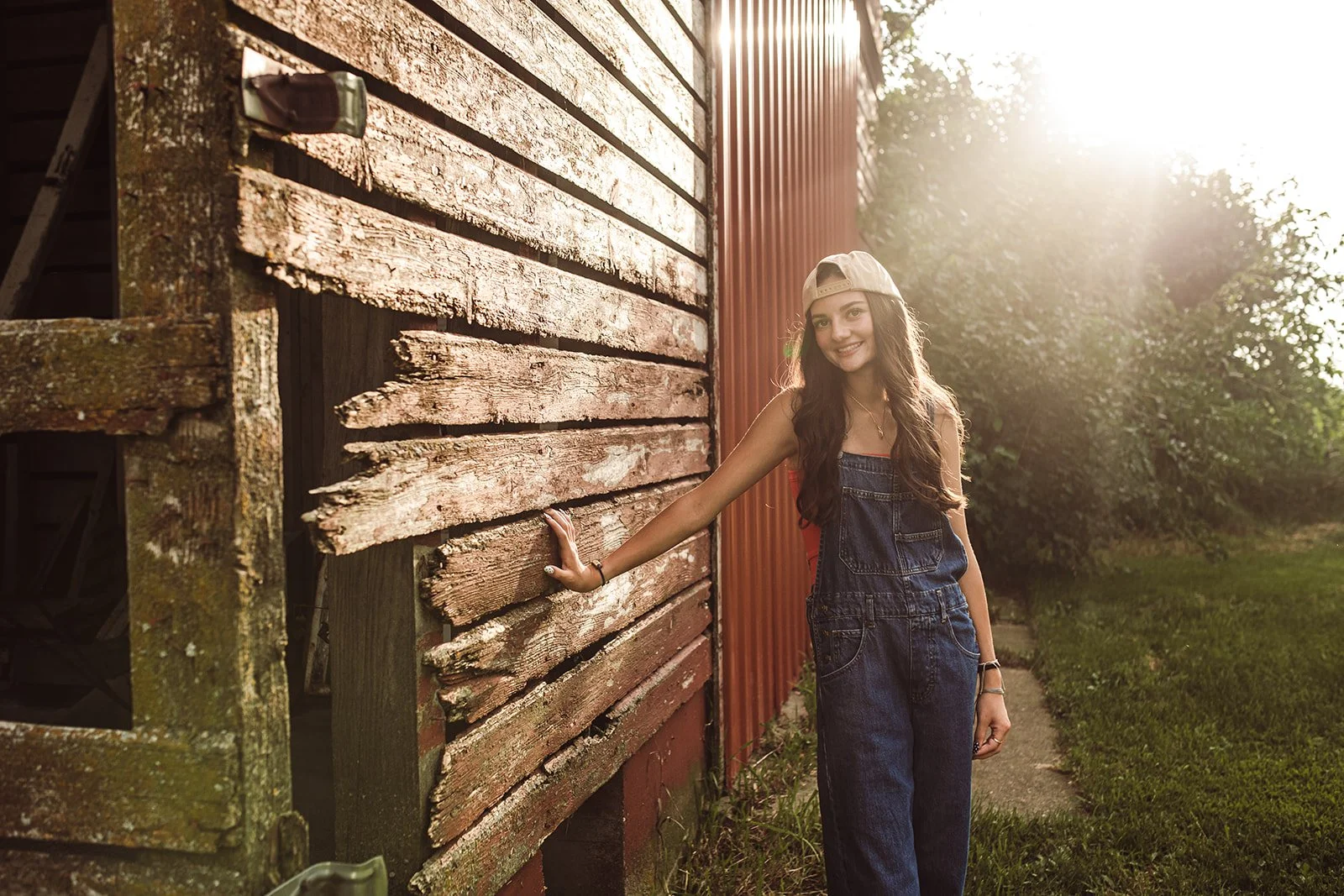 A young woman with long brown hair wearing a beige baseball cap, red shirt, and denim overalls stands outdoors next to a weathered wooden building. She is smiling and leaning against the building on her right hand, with the sunlight shining brightly 