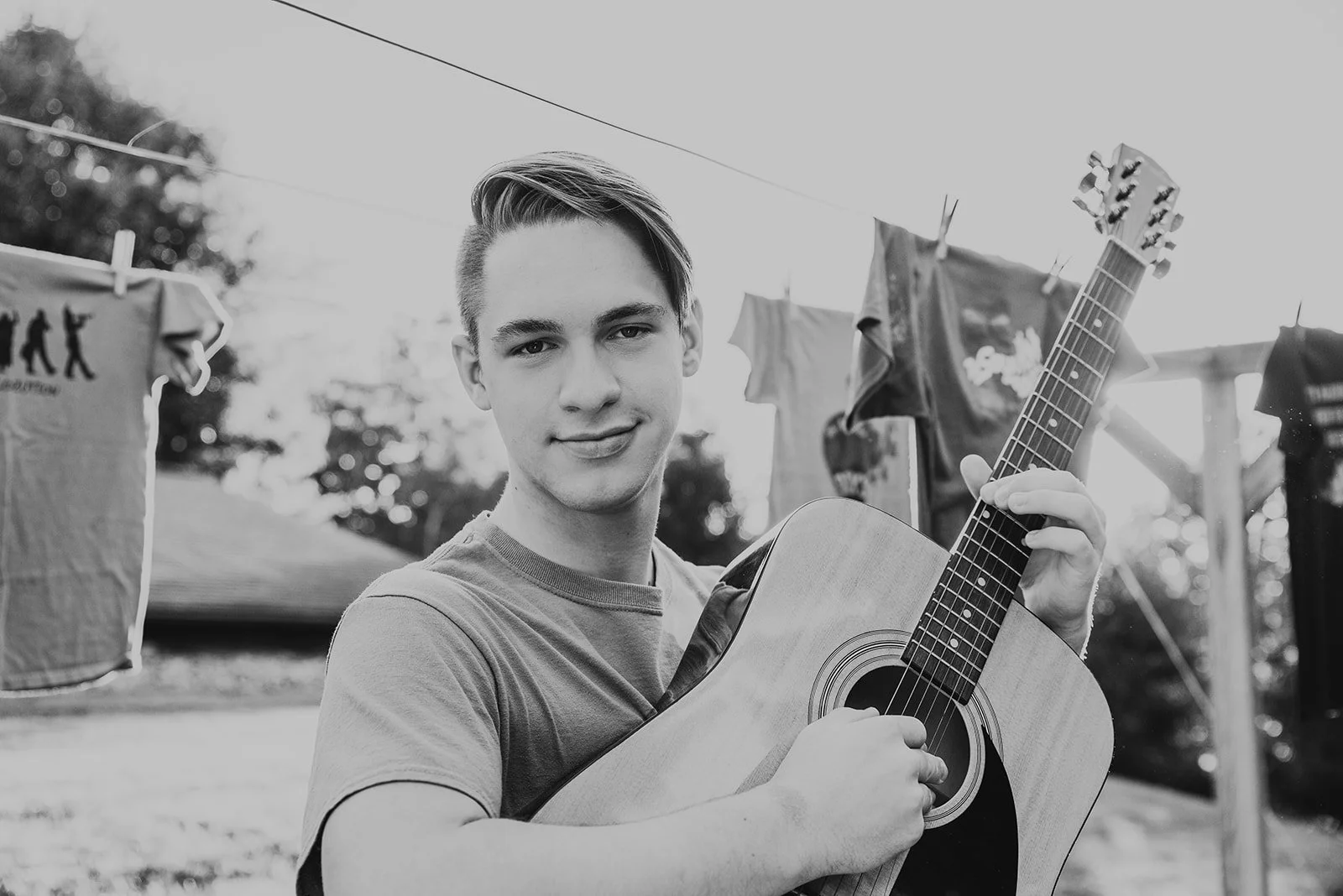 A young man with short, styled hair smiling while holding an acoustic guitar outdoors, with clothes hanging on a line behind him.
