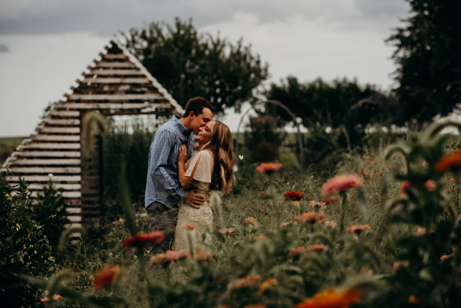 A couple embraces and shares a kiss in a field of wildflowers with a small wooden barn in the background on a cloudy day.