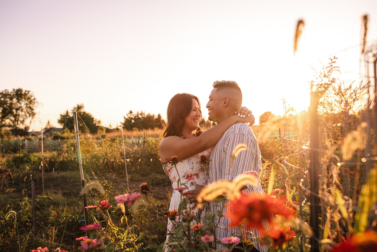 A couple embraces and smiles at each other in a flower garden during sunset, surrounded by blooming flowers and greenery.