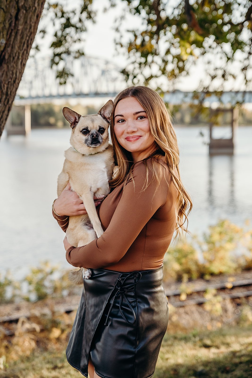 A smiling young woman with long, wavy brown hair holding a small tan Chihuahua with a black nose, near a body of water with a bridge in the background.
