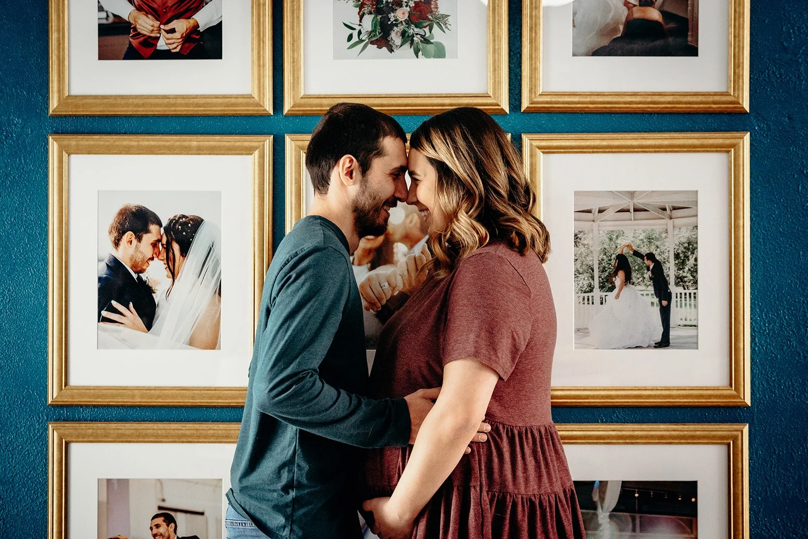 A couple with foreheads touching and smiling faces, standing close in front of a wall with framed wedding photos.