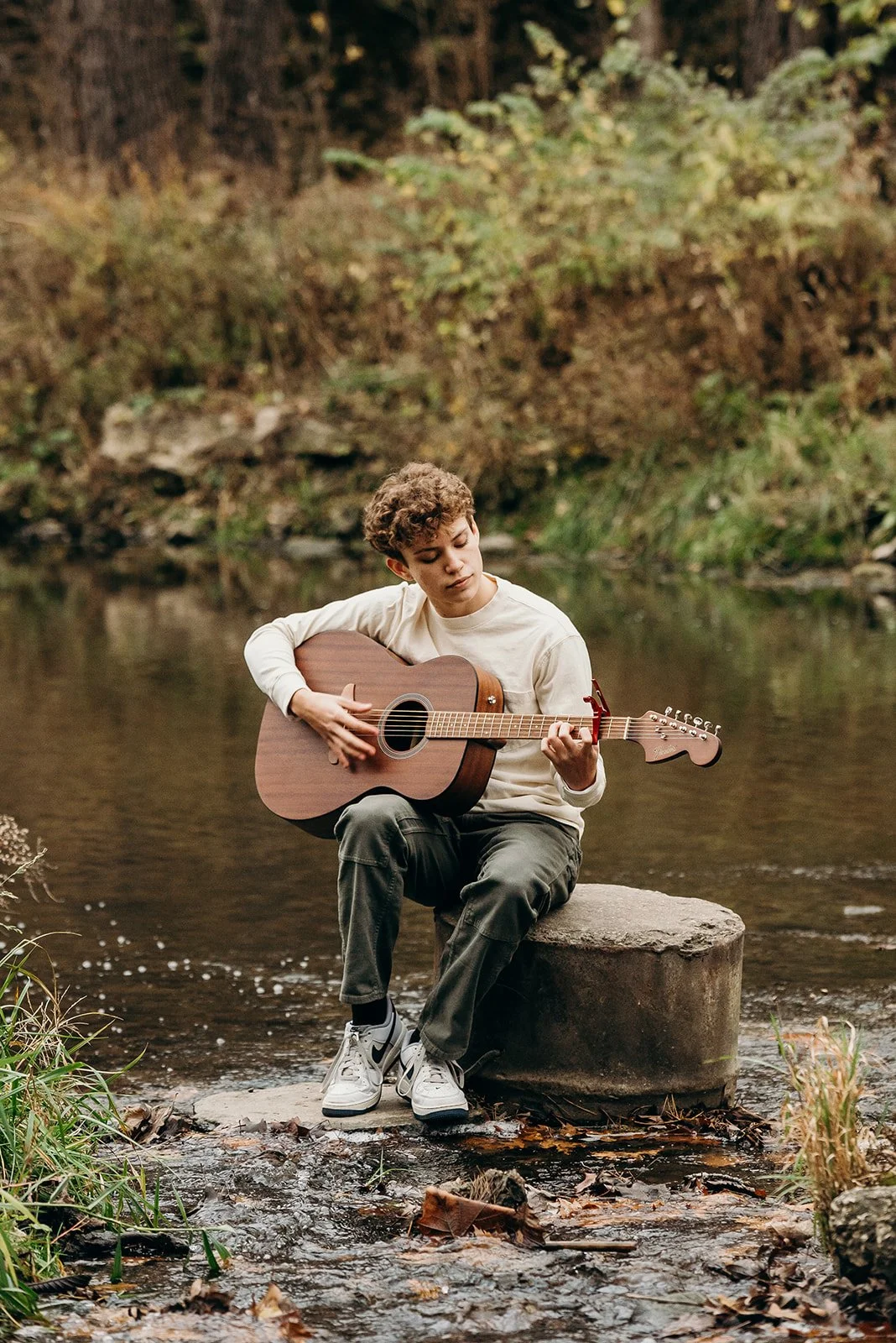 A young man sitting on a concrete block in a creek, playing an acoustic guitar with a background of trees and foliage.
