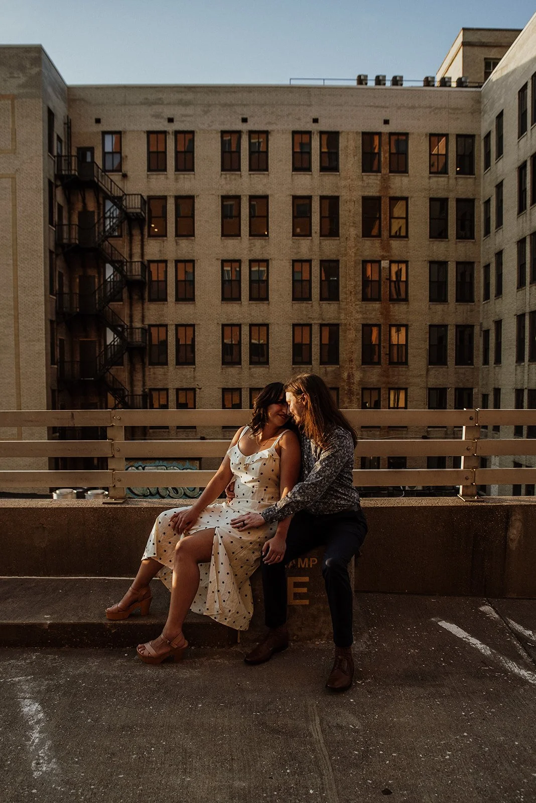 Two women sitting close together on a ledge of a parking garage at sunset, sharing a tender moment, with a large brick apartment building in the background.