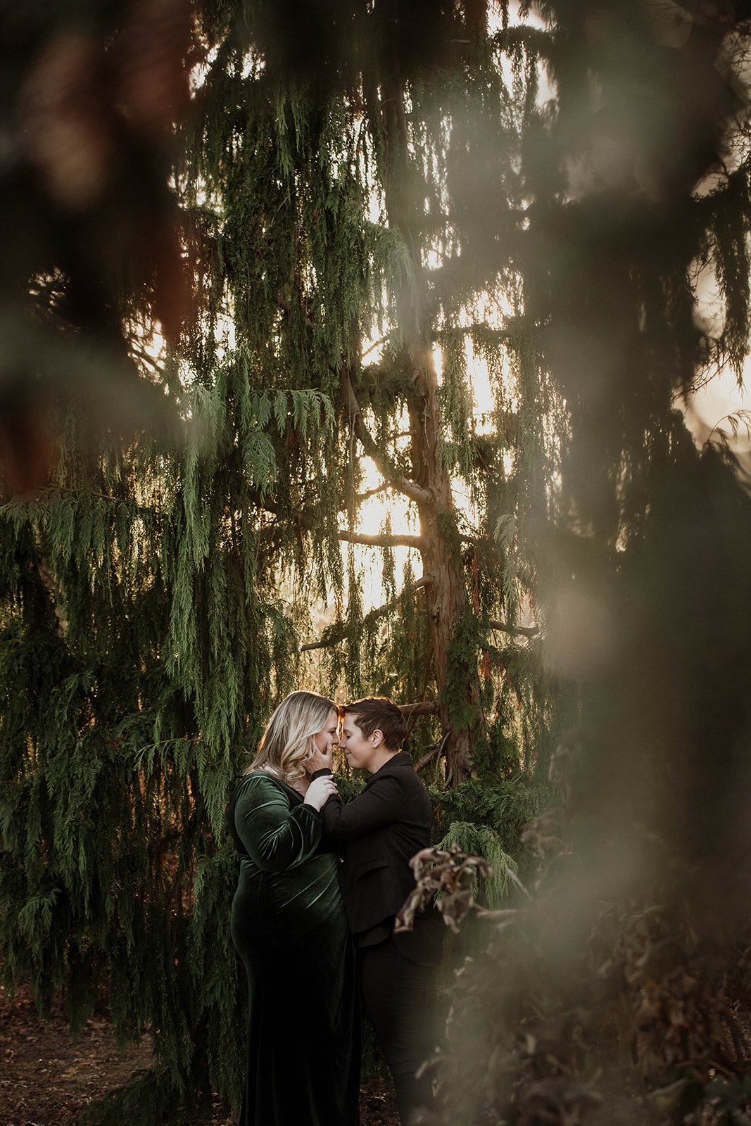 Two women standing close together in front of tall trees, gently touching foreheads with eyes closed during sunset or sunrise.