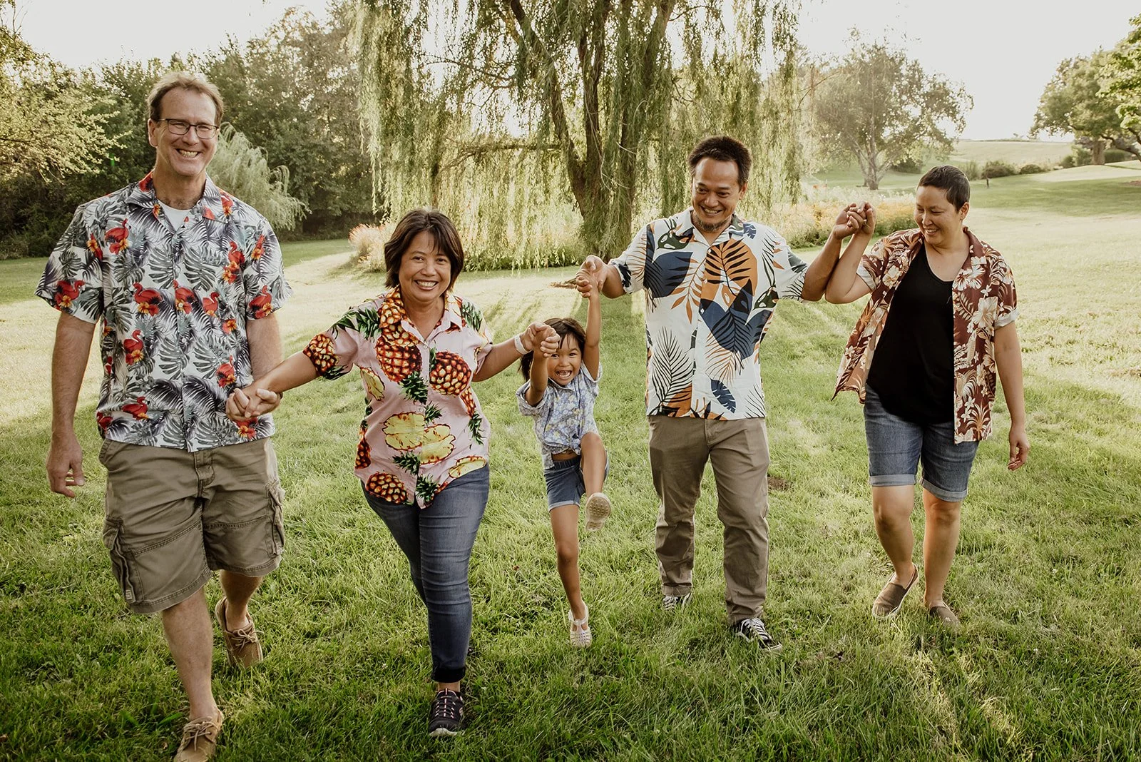 A diverse group of five people, including two men, two women, and a young girl, holding hands and walking on a lush green park, smiling and enjoying a sunny day.