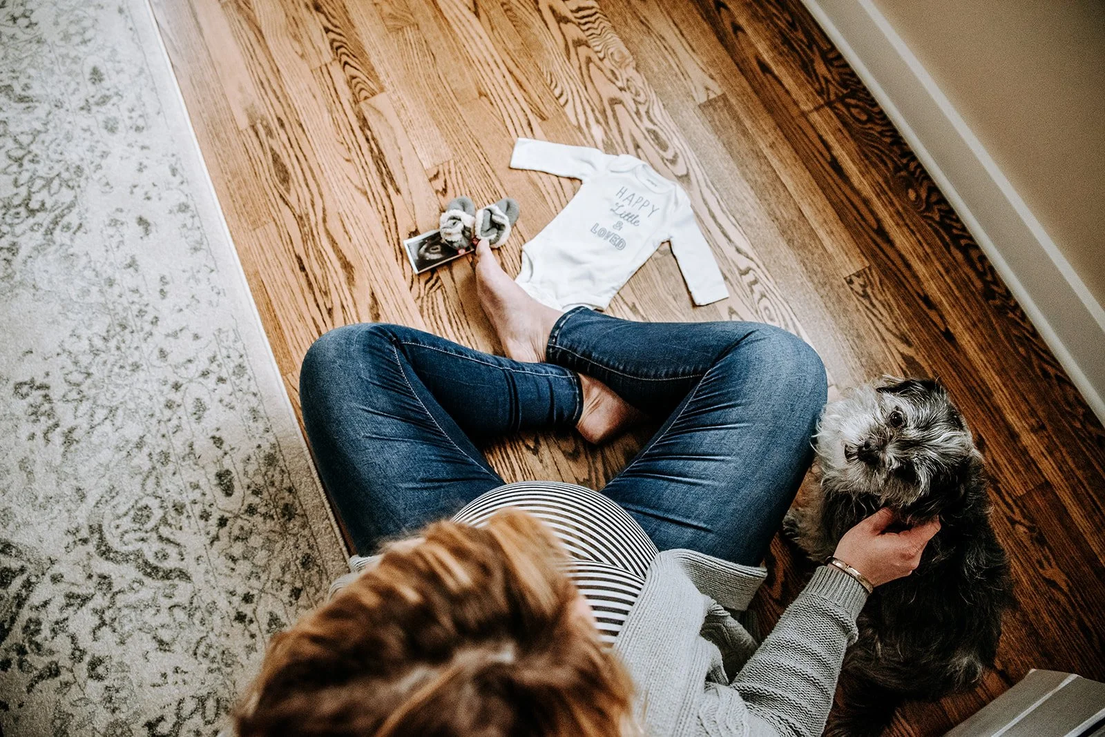 A person sitting on the floor with crossed legs, petting a small dog while another dog is near her feet. On the floor, there is a white bib with "HAPPY Little & LOUD" printed on it, a photo, and a pair of slippers.