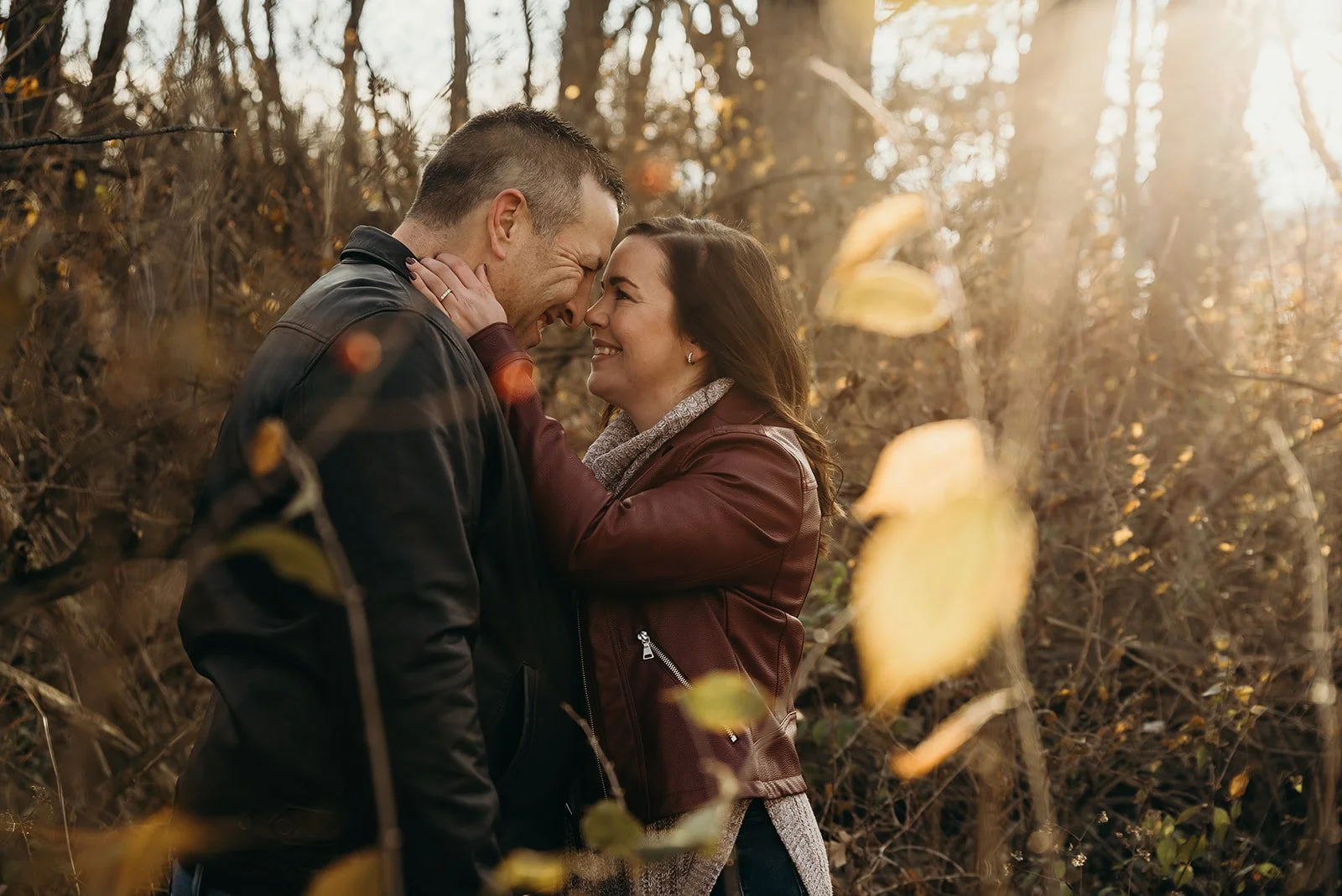 A couple in an outdoor fall setting with trees and leaves, smiling and touching foreheads, with warm sunlight shining through the trees.
