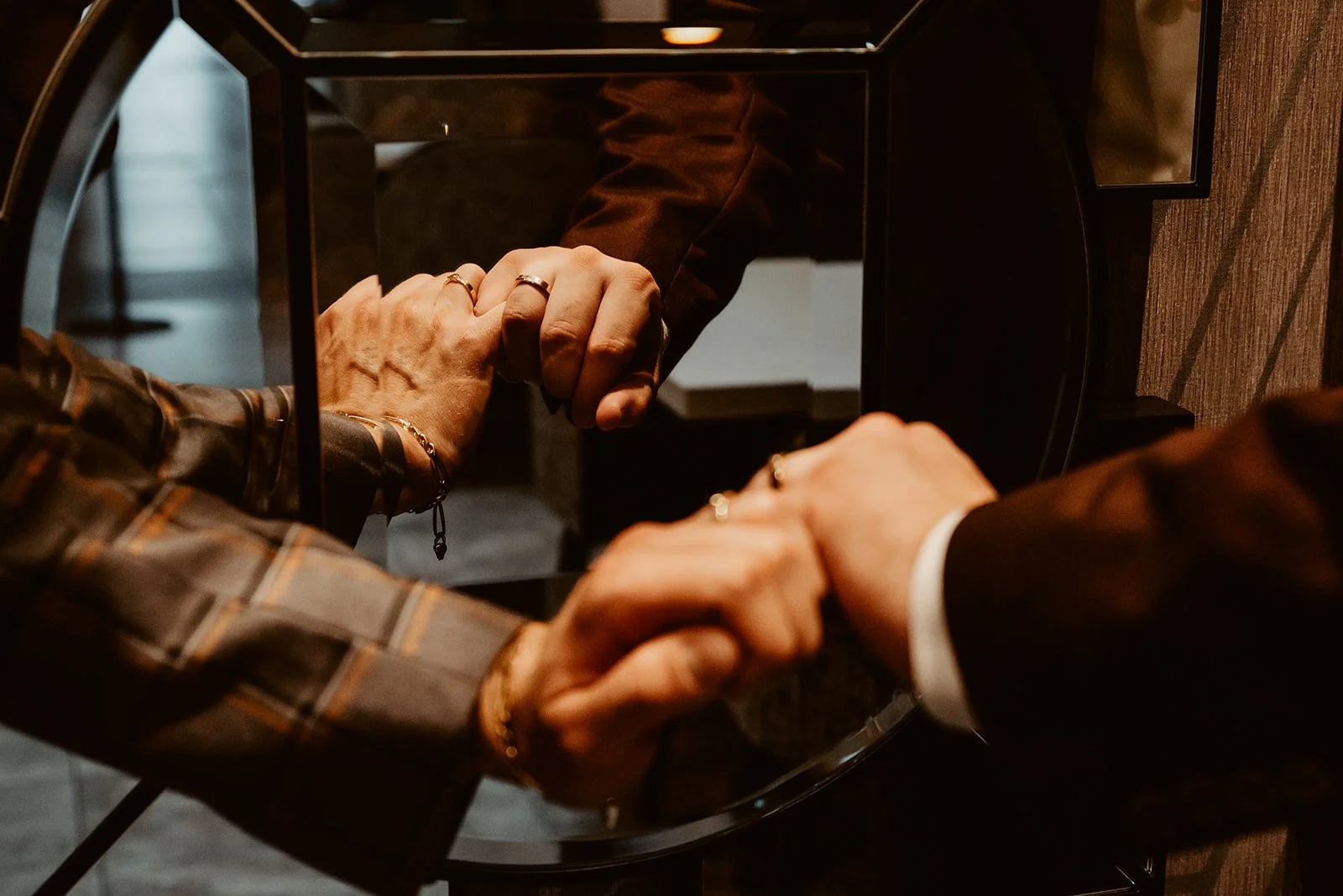 Close-up of two men's hands clasped together in front of a reflective mirror, showing rings and jewelry, with a dark setting.
