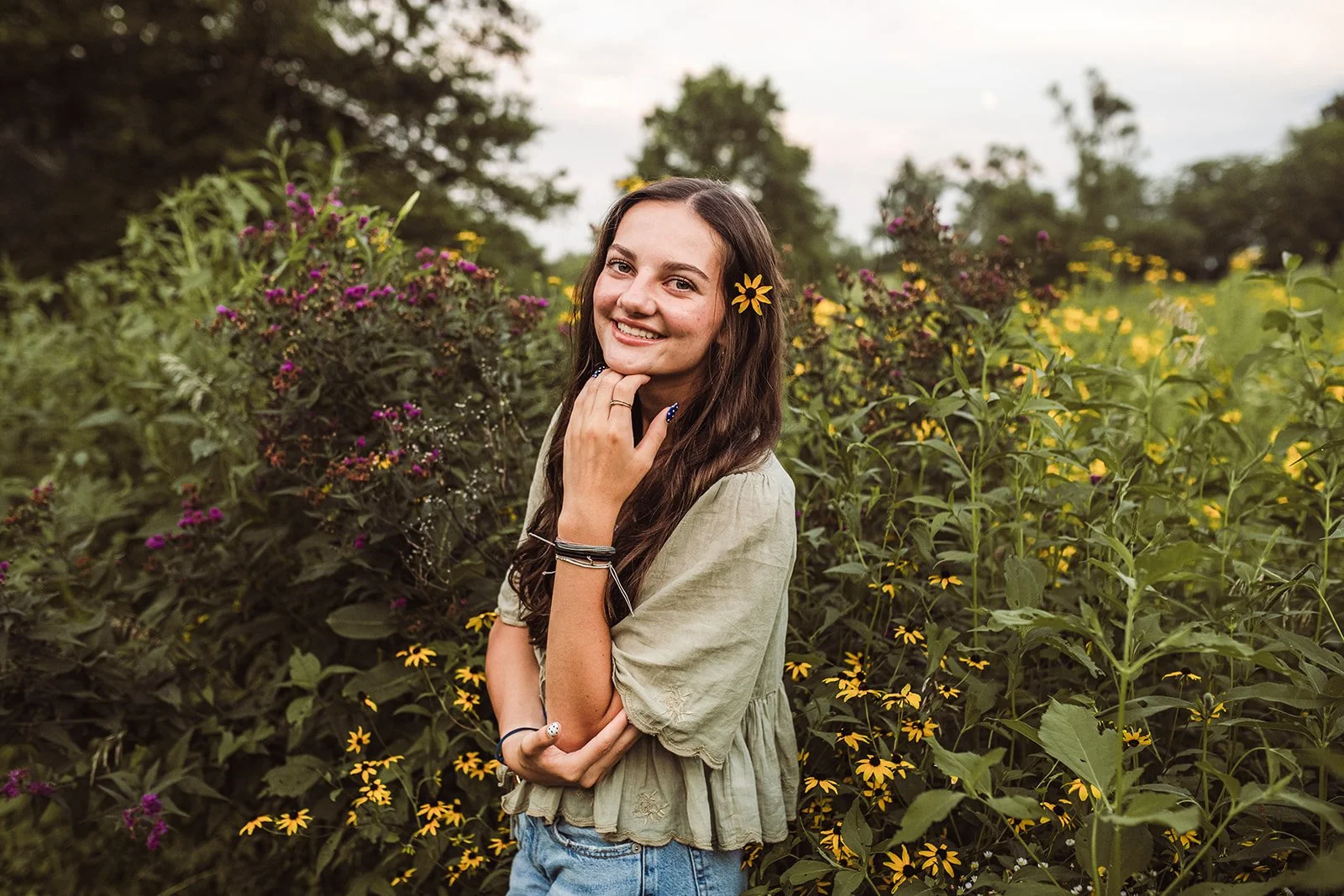 Young woman with long brown hair smiling in a field of tall yellow and purple flowers, wearing a light green blouse, with a flower tucked in her hair.