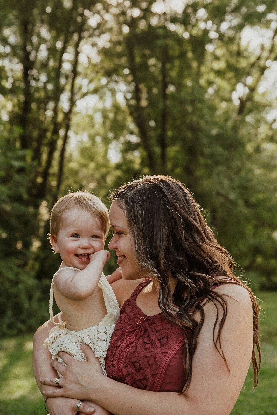 A woman holding a smiling toddler outdoors in a wooded area, both enjoying a moment of happiness.