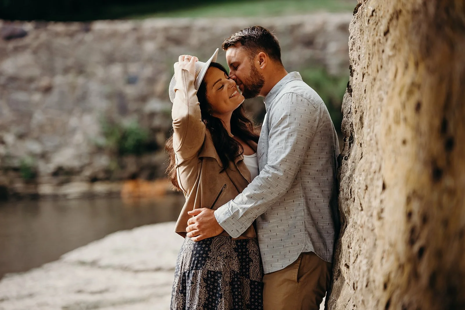 A couple sharing an intimate moment by a river, with the woman holding a hat and smiling, and the man leaning in toward her as they embrace.