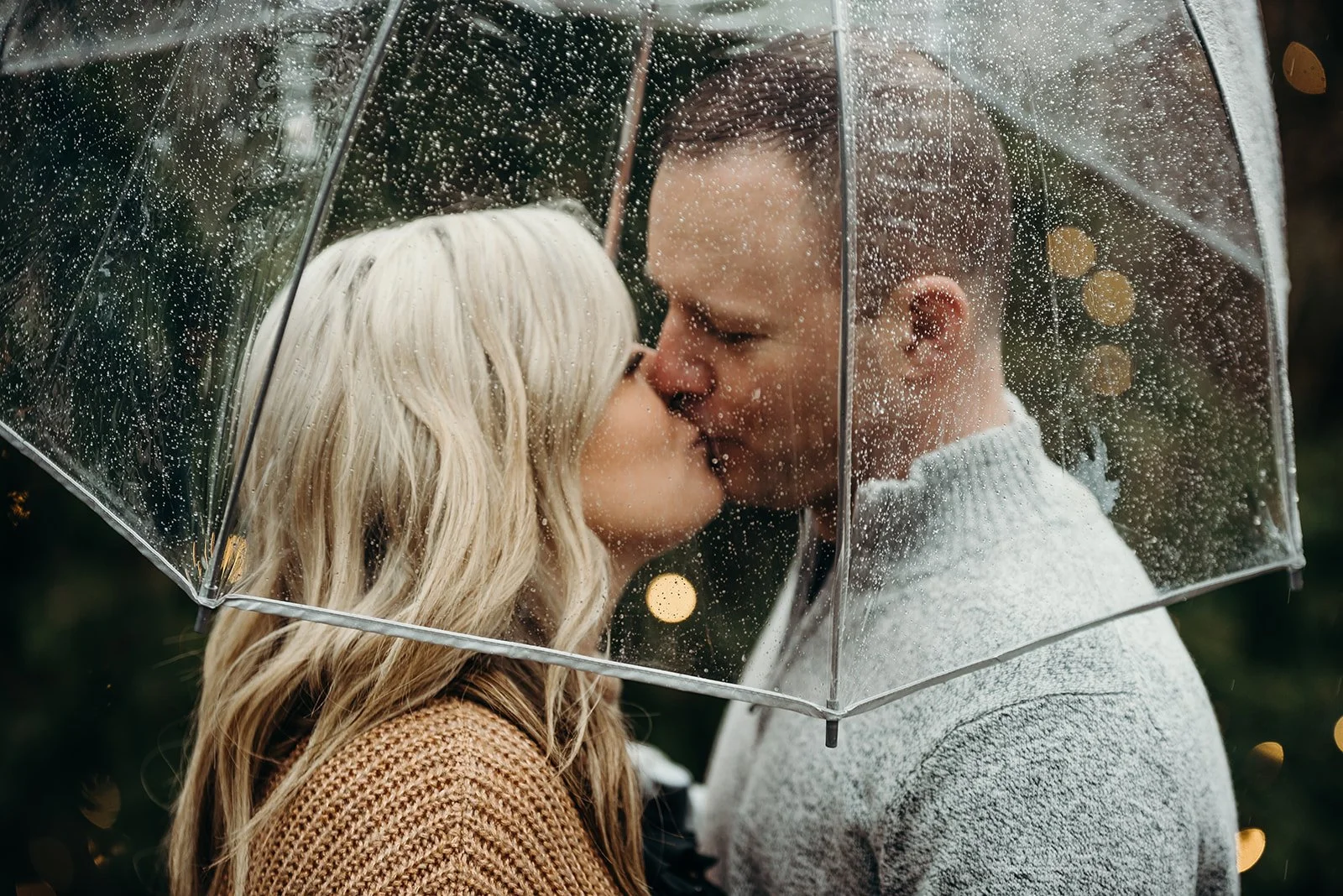 A couple sharing a kiss under a transparent umbrella with rain droplets, outdoors in a blurred background.