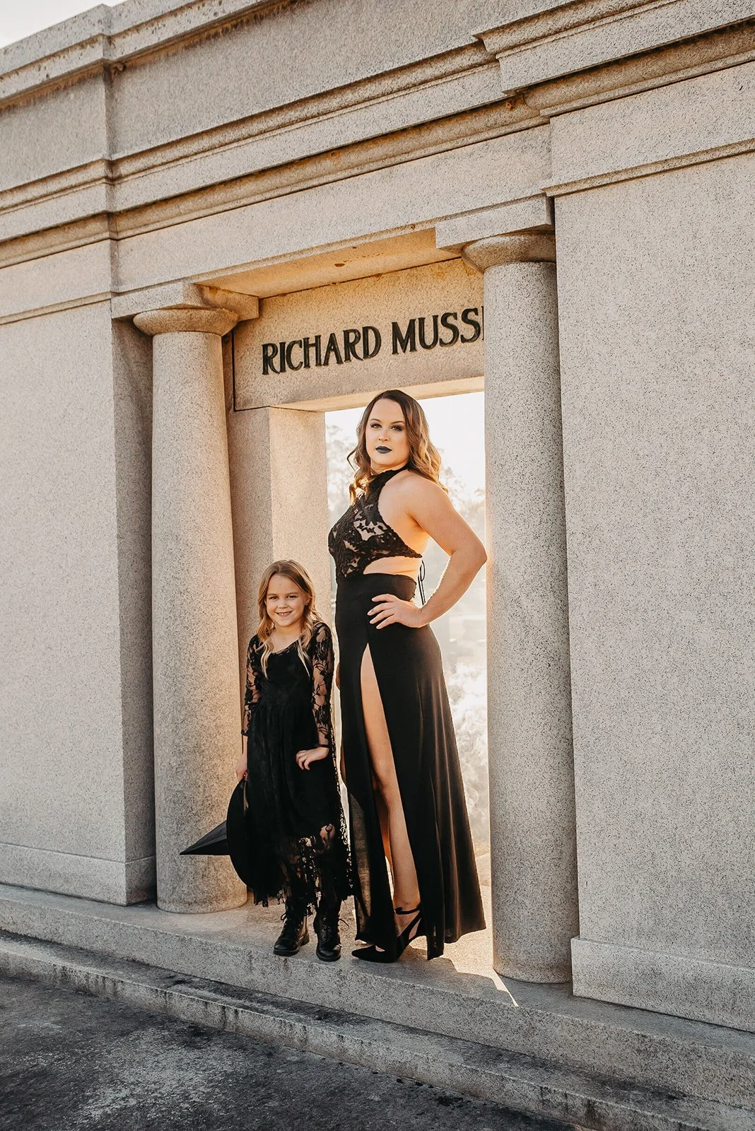 A woman and a young girl dressed in black evening gowns standing under a stone arch with the words 'Richard Mussa' on it. The woman is taller, with shoulder-length wavy hair and dark makeup, and the girl has long, blonde hair and a big smile.